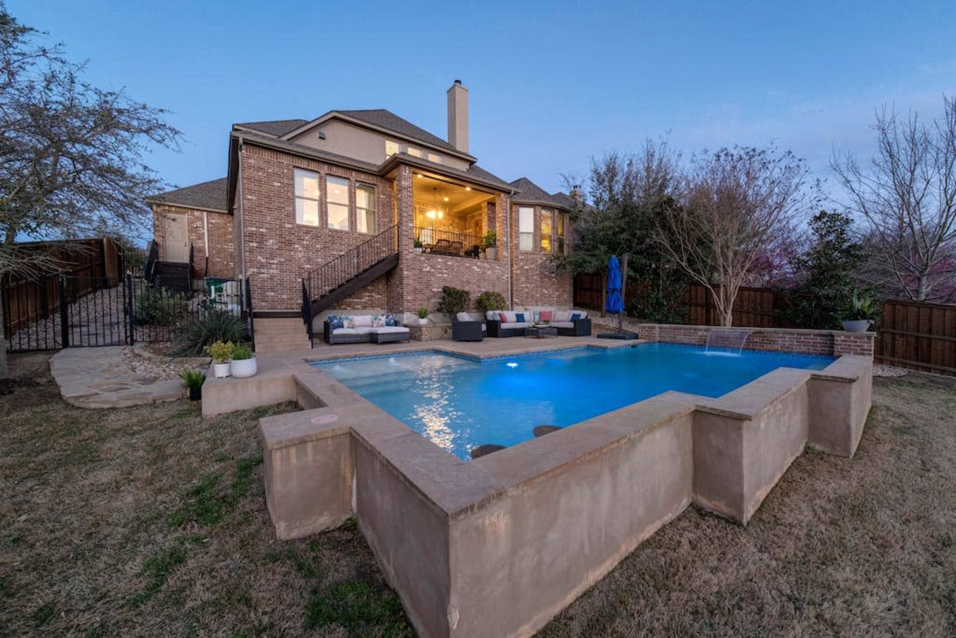 A backyard features a rectangular swimming pool with a fountain, surrounded by grass and stone landscaping, alongside a two-story brick house with outdoor seating.