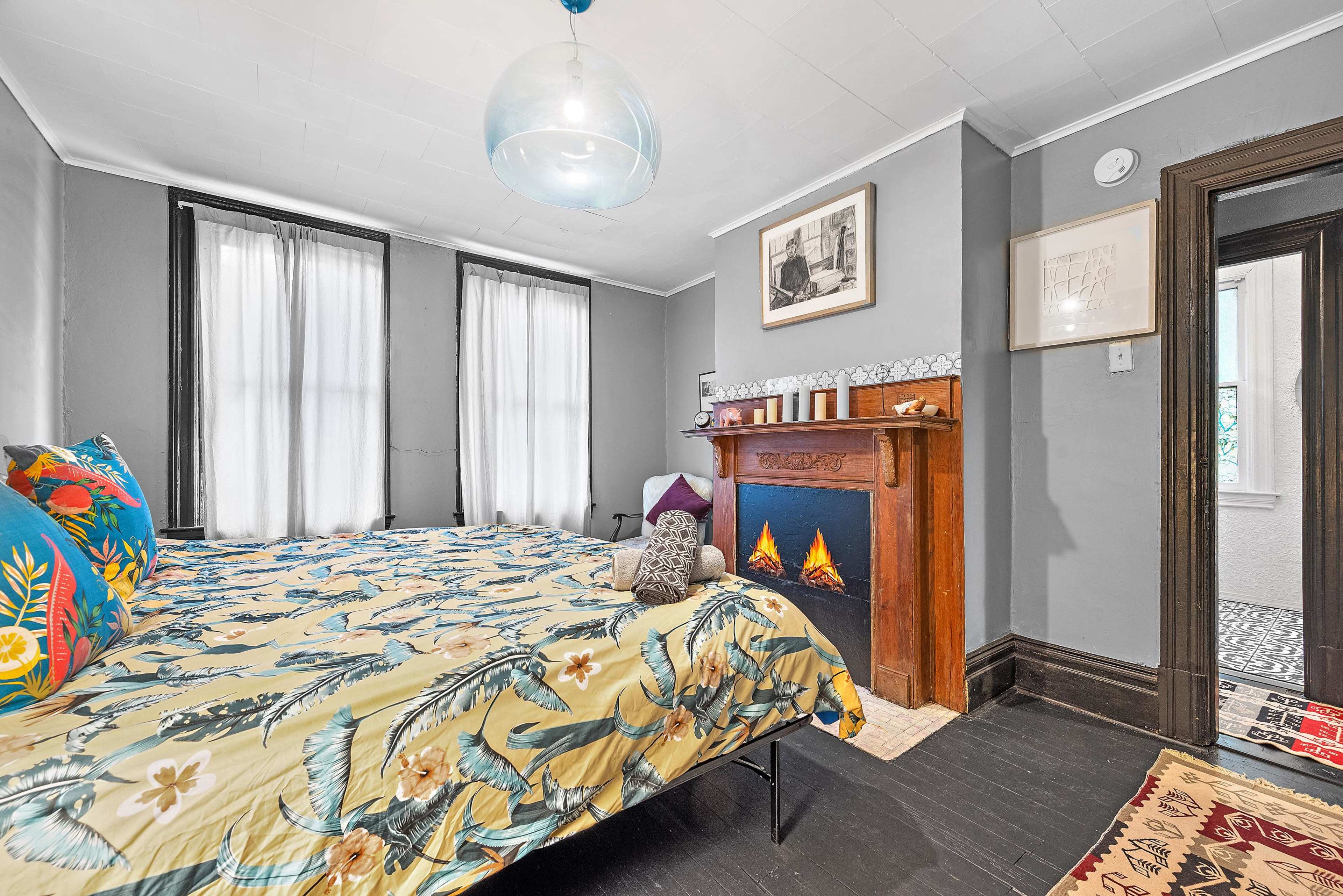 A bedroom with a patterned bedspread, two large windows with sheer curtains, and a decorative fireplace surrounded by candles and artwork.