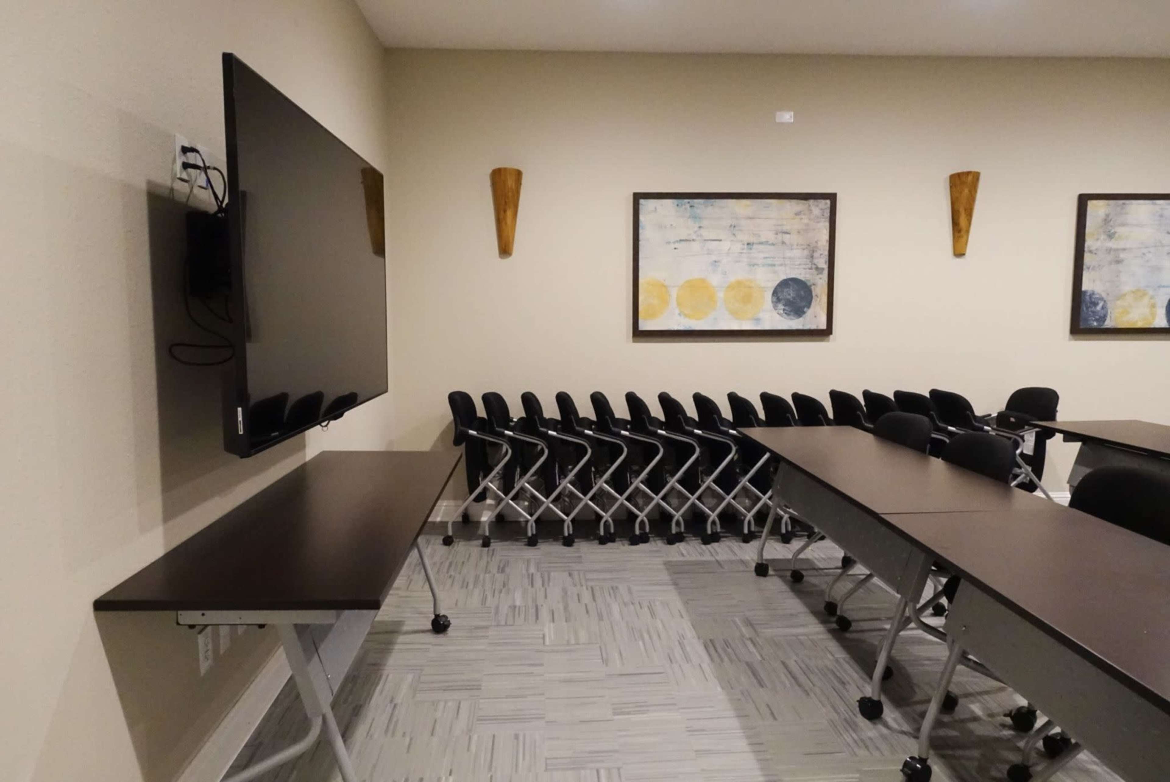 A conference room features a large television mounted on the wall, with rows of stacked chairs and tables arranged in the space.