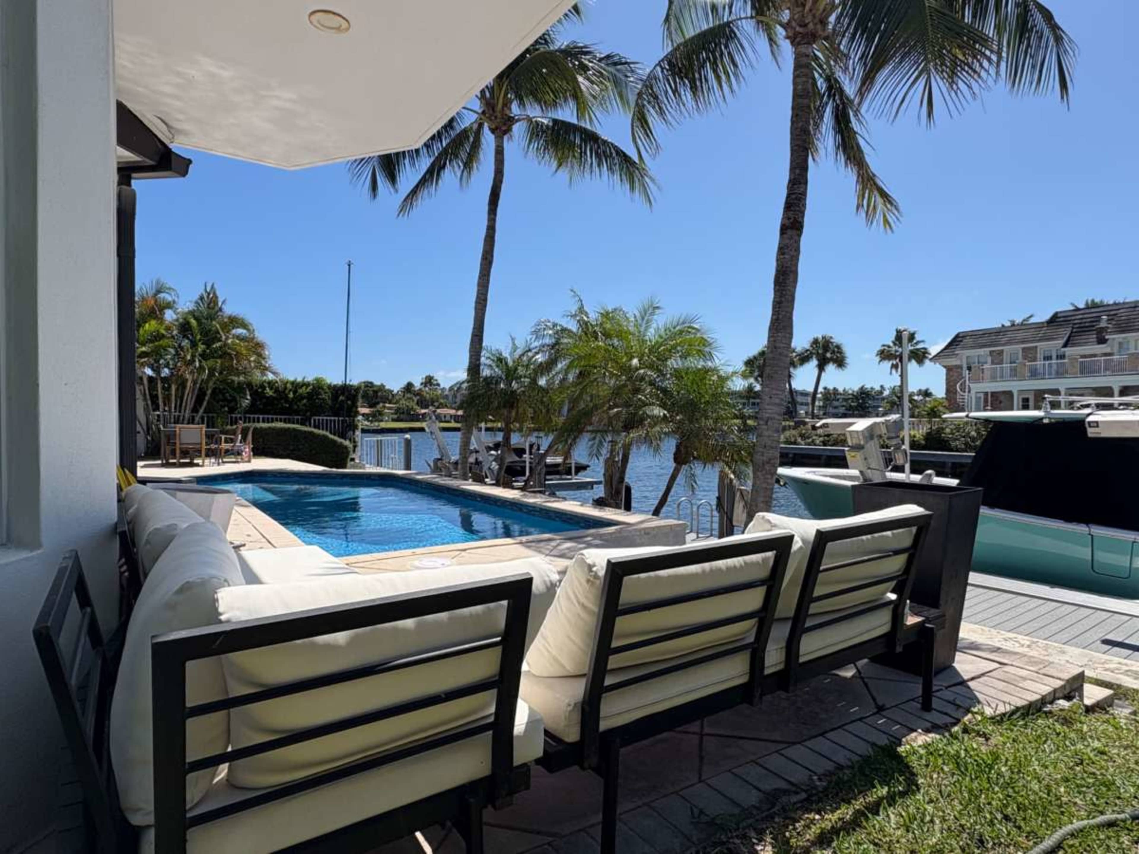 The image shows a backyard scene with a swimming pool, palm trees, and a waterway, along with lounge seating facing the water.