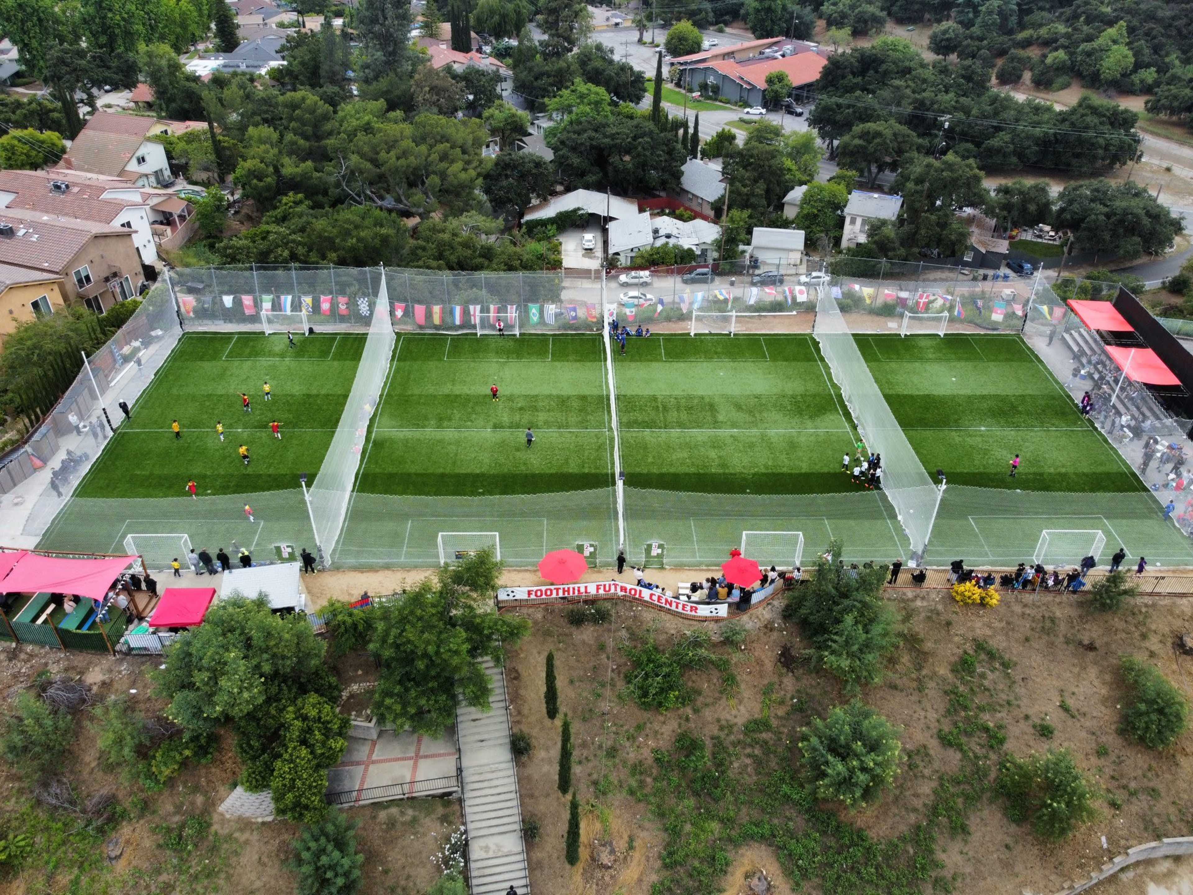 The image shows an aerial view of two soccer fields with players engaged in matches, surrounded by trees and residential buildings.