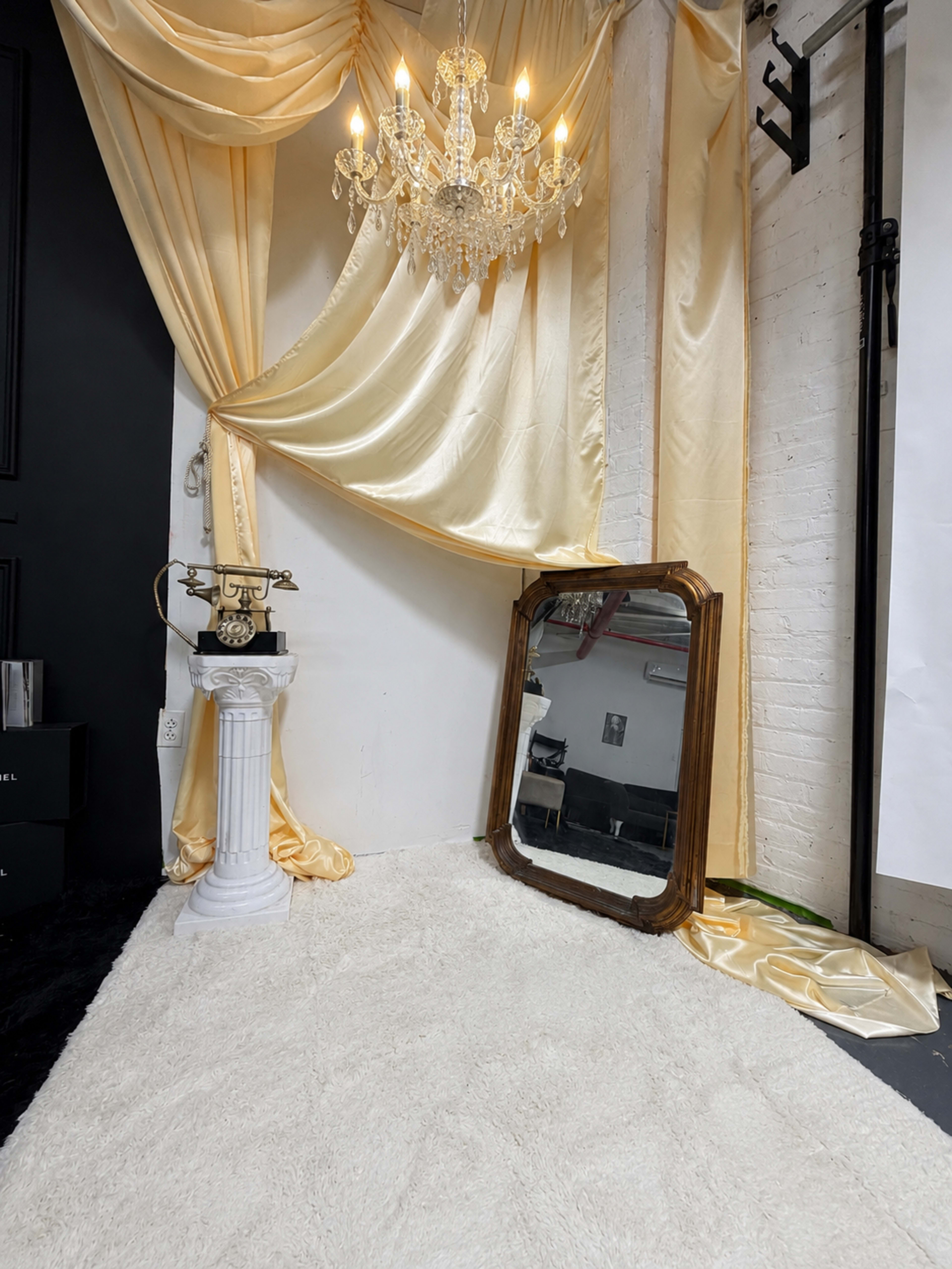 The scene features a corner with a vintage telephone on a white pedestal, a large ornate mirror leaning against a wall, and a crystal chandelier hanging above, all set against a backdrop of draped yellow fabric.