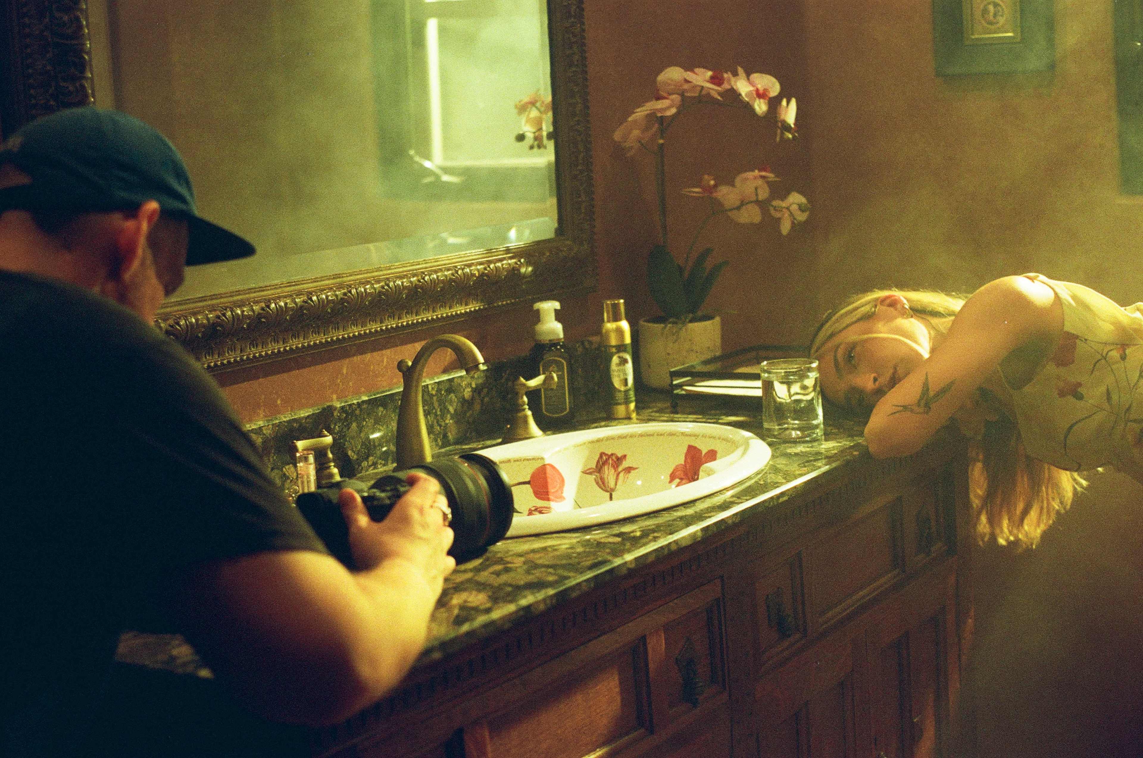 A photographer captures an image of a woman resting her head on a bathroom counter next to a sink adorned with a floral-patterned bowl.