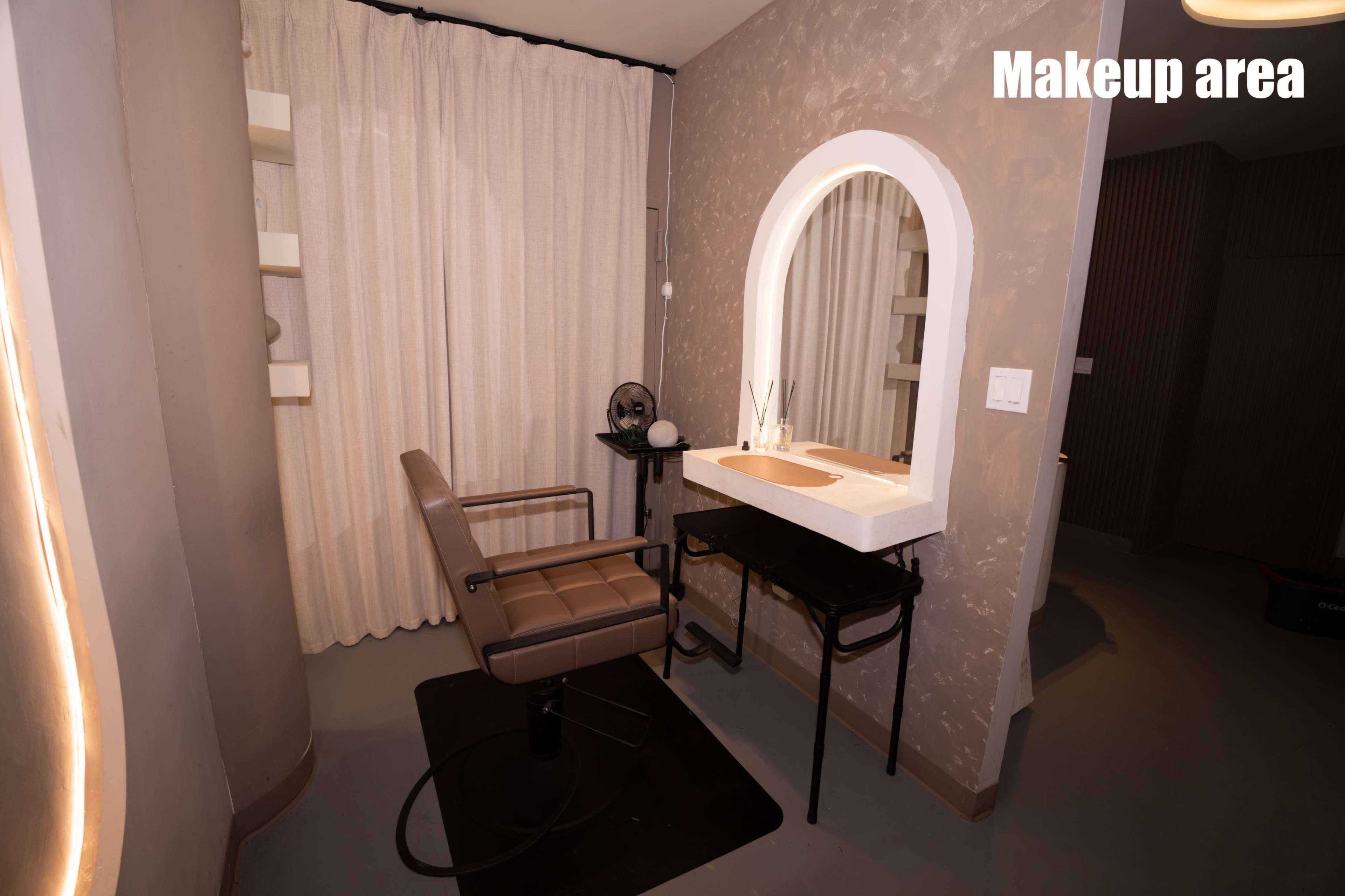 The image shows a makeup area featuring a circular mirror above a countertop with a chair beside it, surrounded by soft curtain drapes.