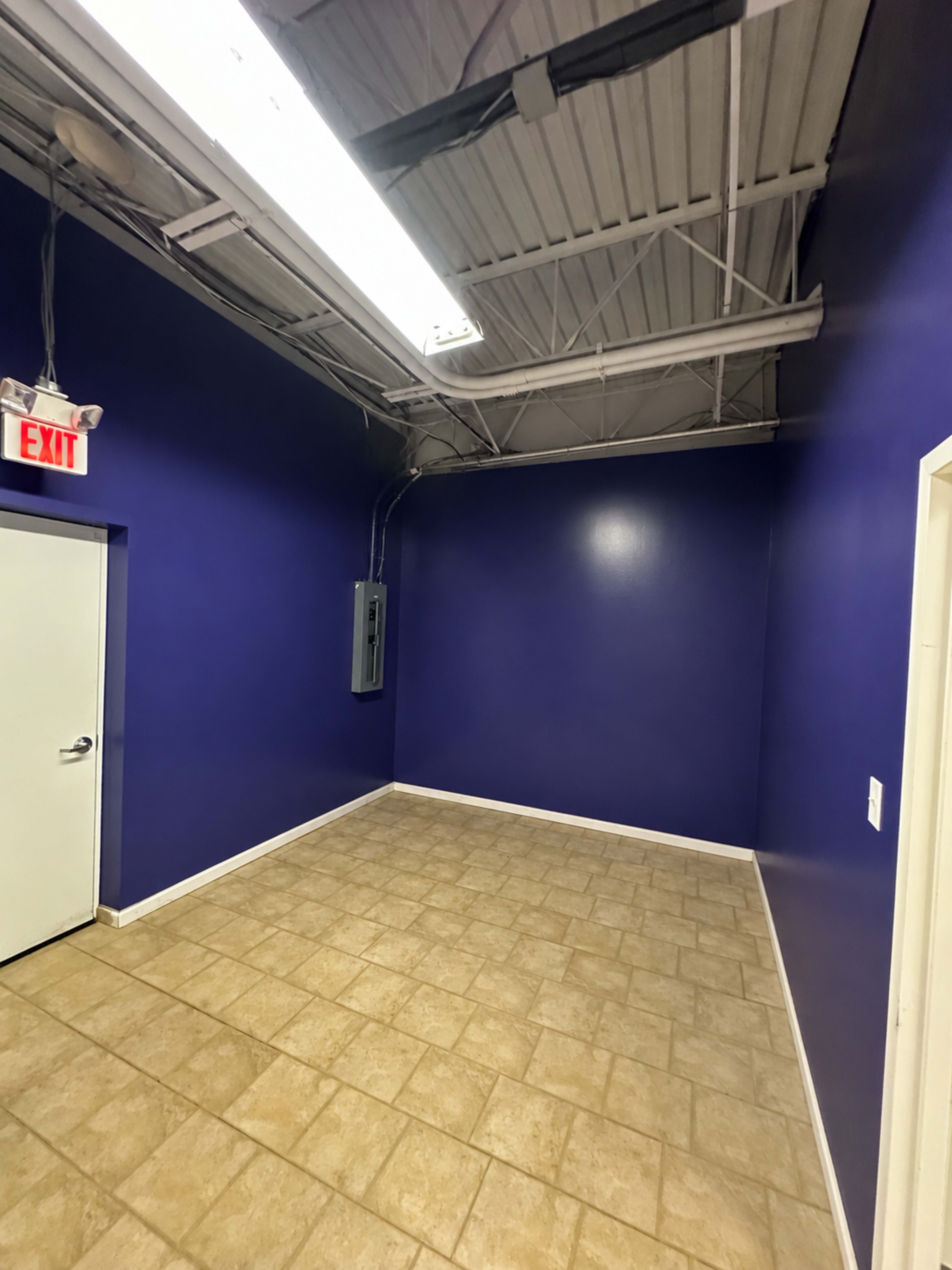 The image shows a vacant room with purple walls, tiled flooring, a white door, and exposed ceiling infrastructure.