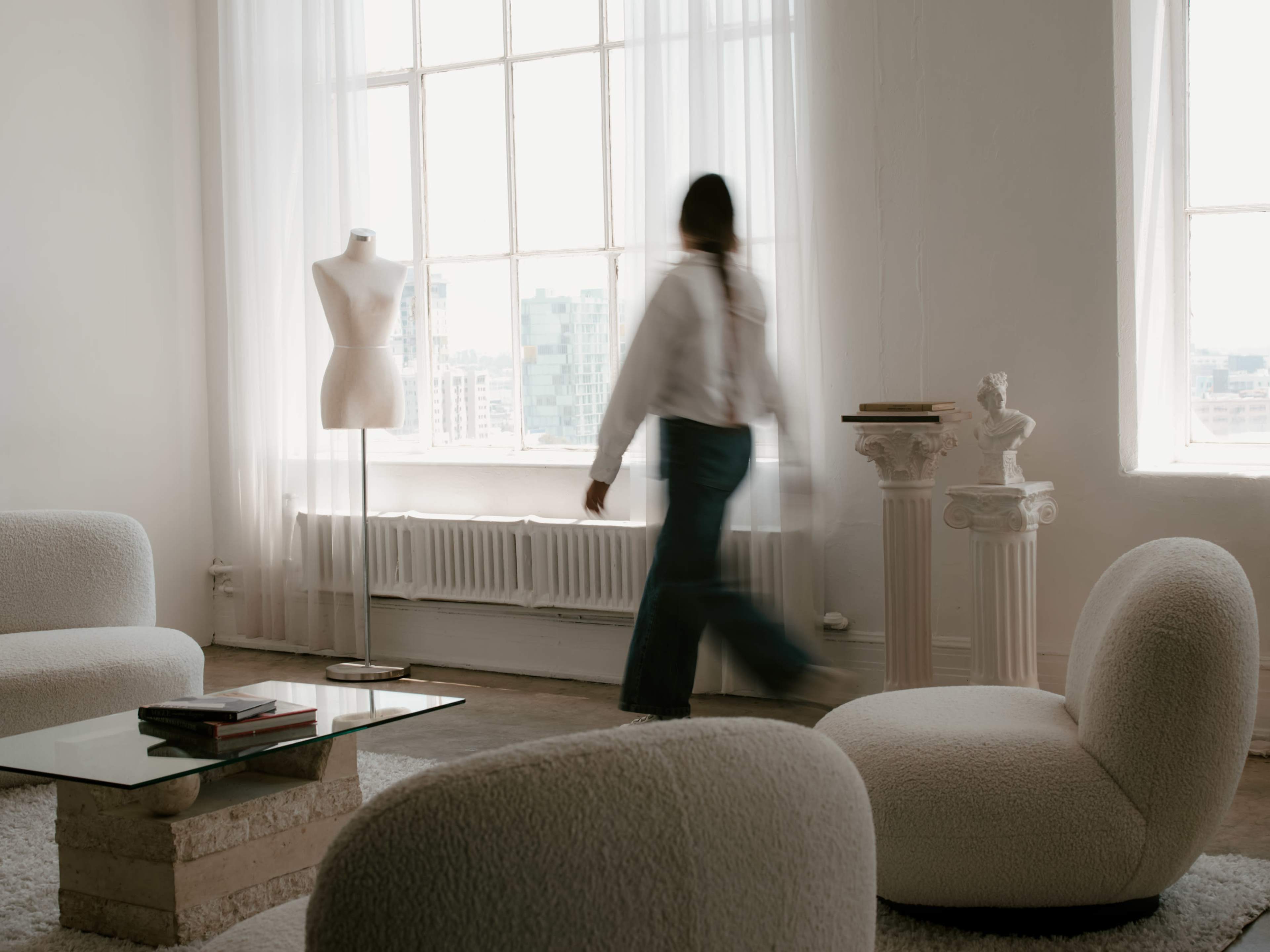 A person walks past a dress form in a minimally furnished room with large windows and soft furniture.