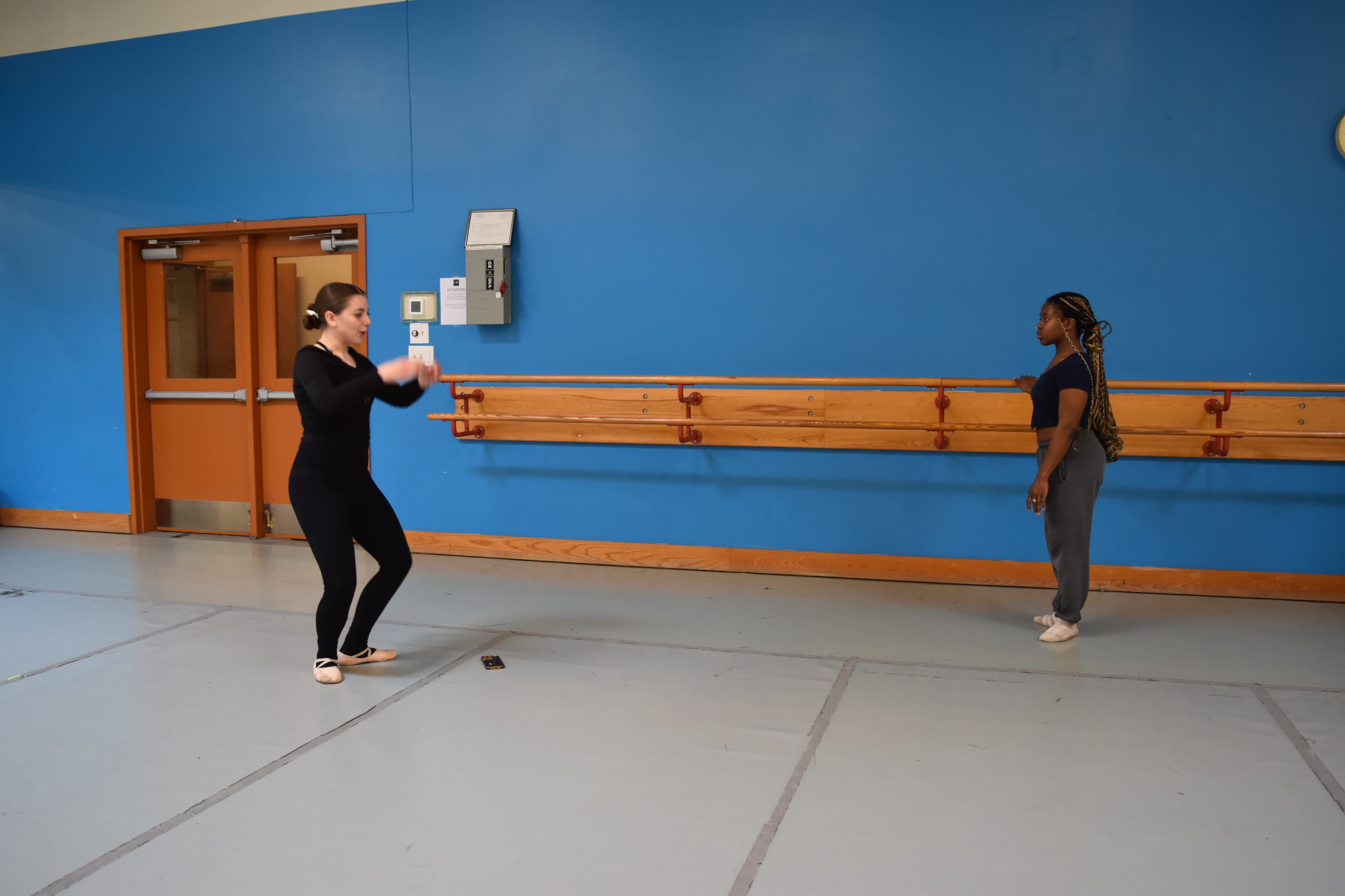 Two dancers practice in a ballet studio with a blue wall and wooden ballet bar.