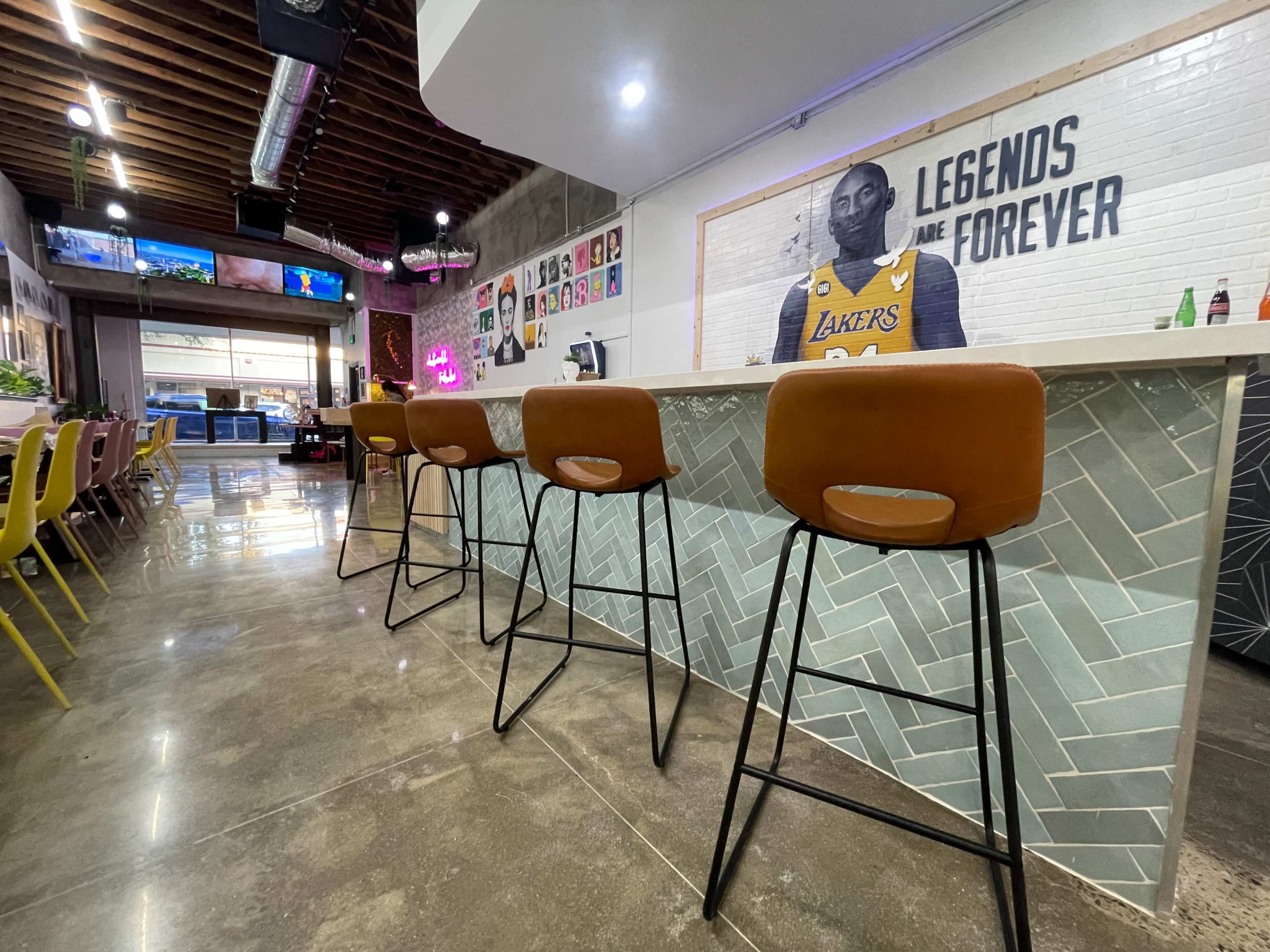 A modern bar area with high stools, a herringbone-patterned counter, and a mural of a basketball player on the wall in a restaurant setting.