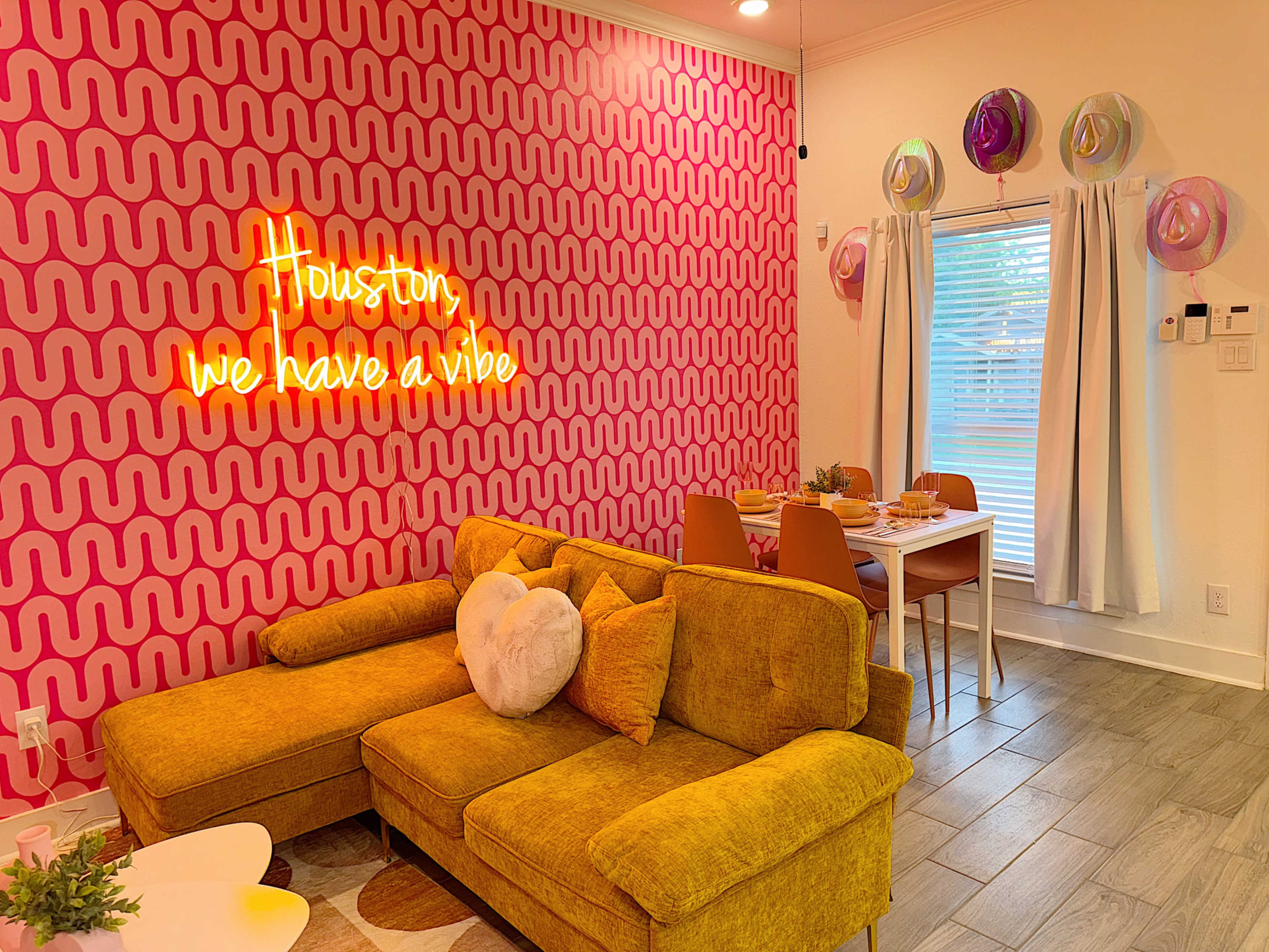 A living room features a yellow sofa, pink wavy-patterned wallpaper, and a neon sign that reads "Houston we have a vibe."