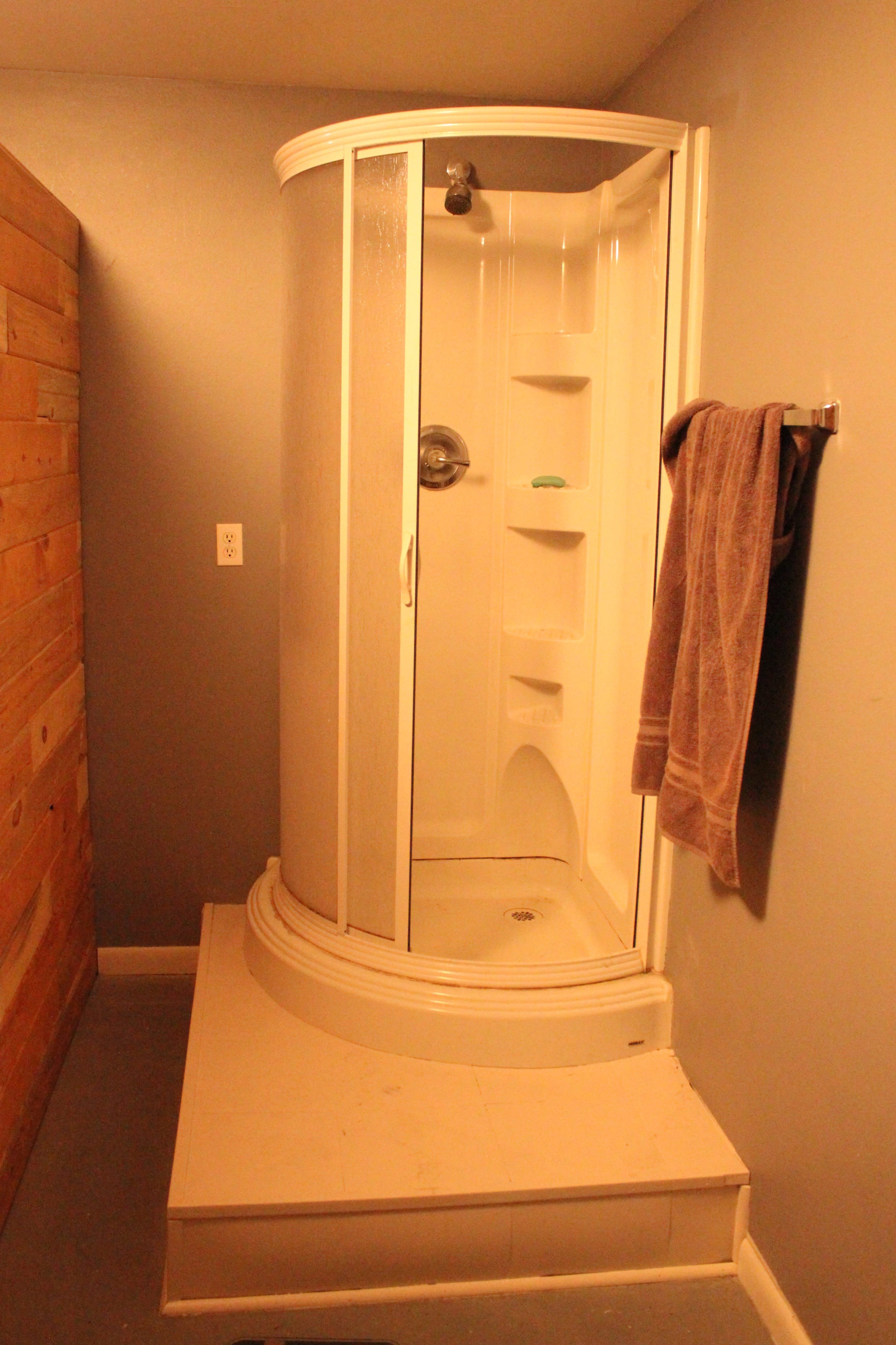 The image shows a corner shower with a curved glass door, situated in a bathroom with wooden paneling on one wall.