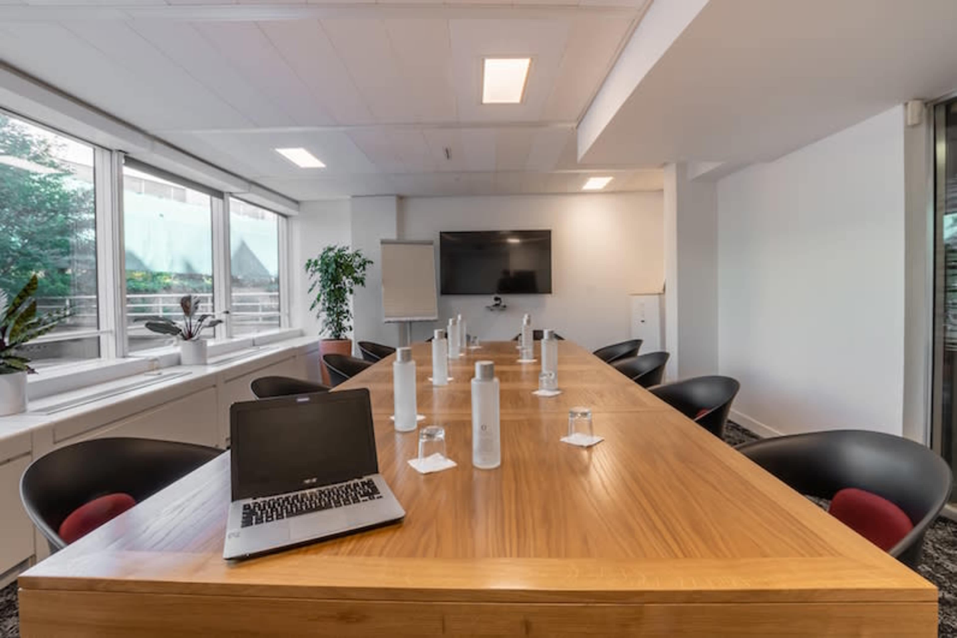 A modern conference room features a long wooden table surrounded by black chairs, with water bottles and glasses set for a meeting.