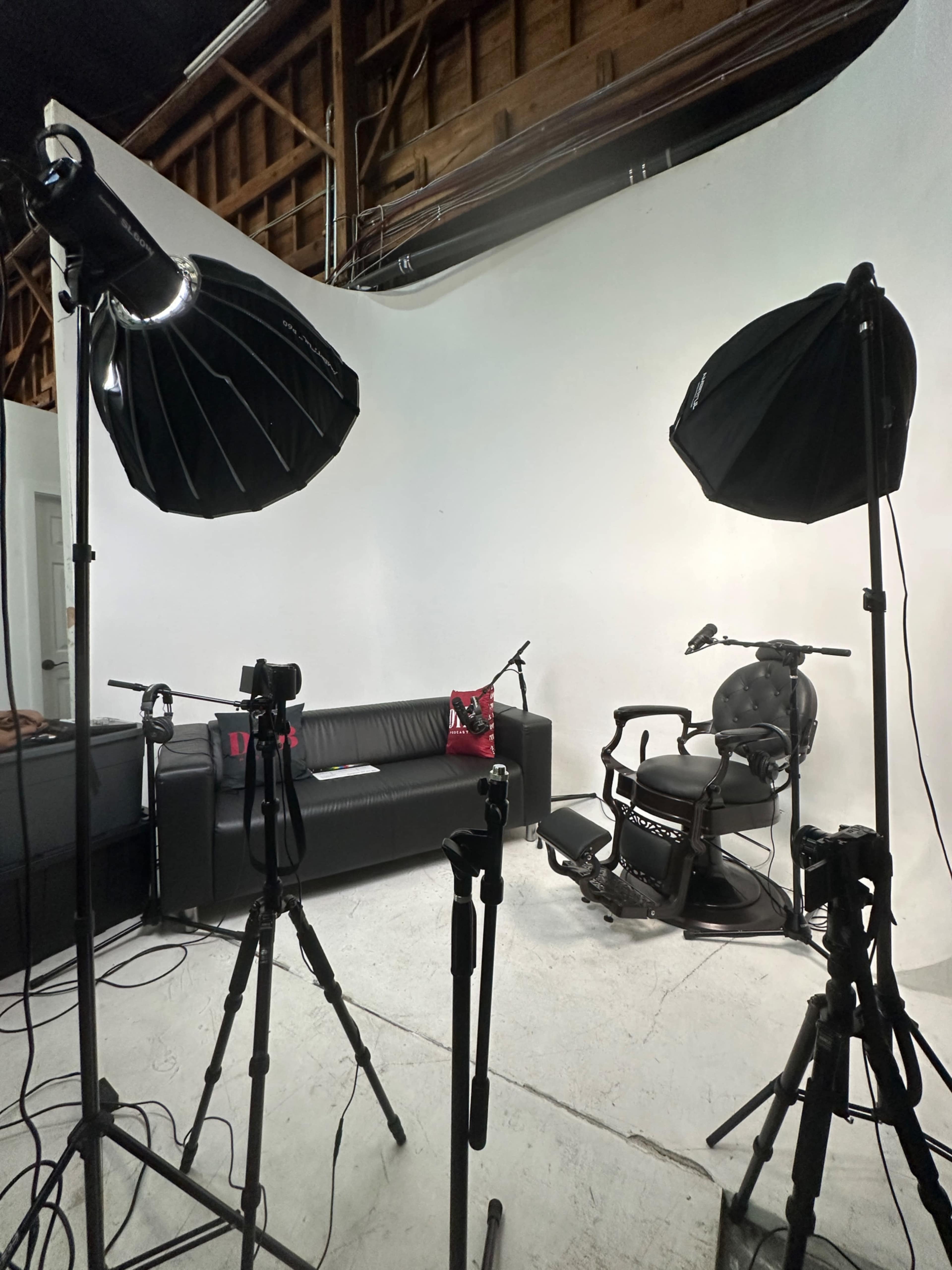 The image shows a photography studio with lighting equipment, a black leather sofa, and a black chair set against a white backdrop.