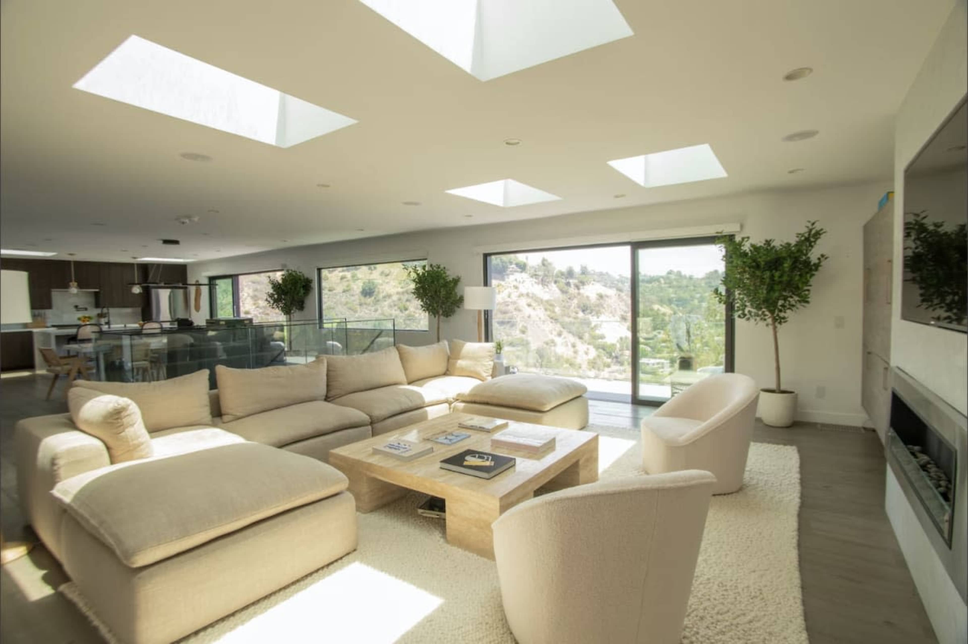 The living room features a large sectional sofa, two armchairs, and a coffee table, with floor-to-ceiling windows providing a view of a hillside.