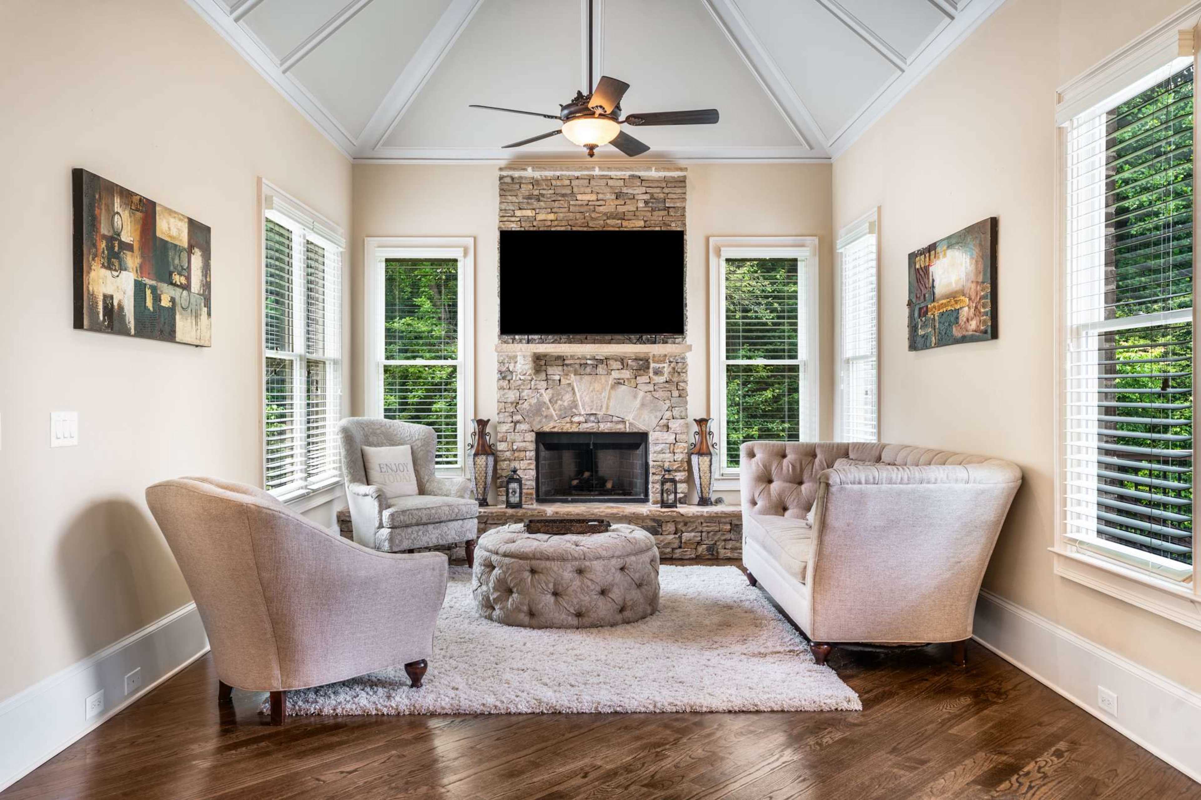 A cozy living room features two sofas, a round ottoman, and a stone fireplace under a vaulted ceiling with large windows.