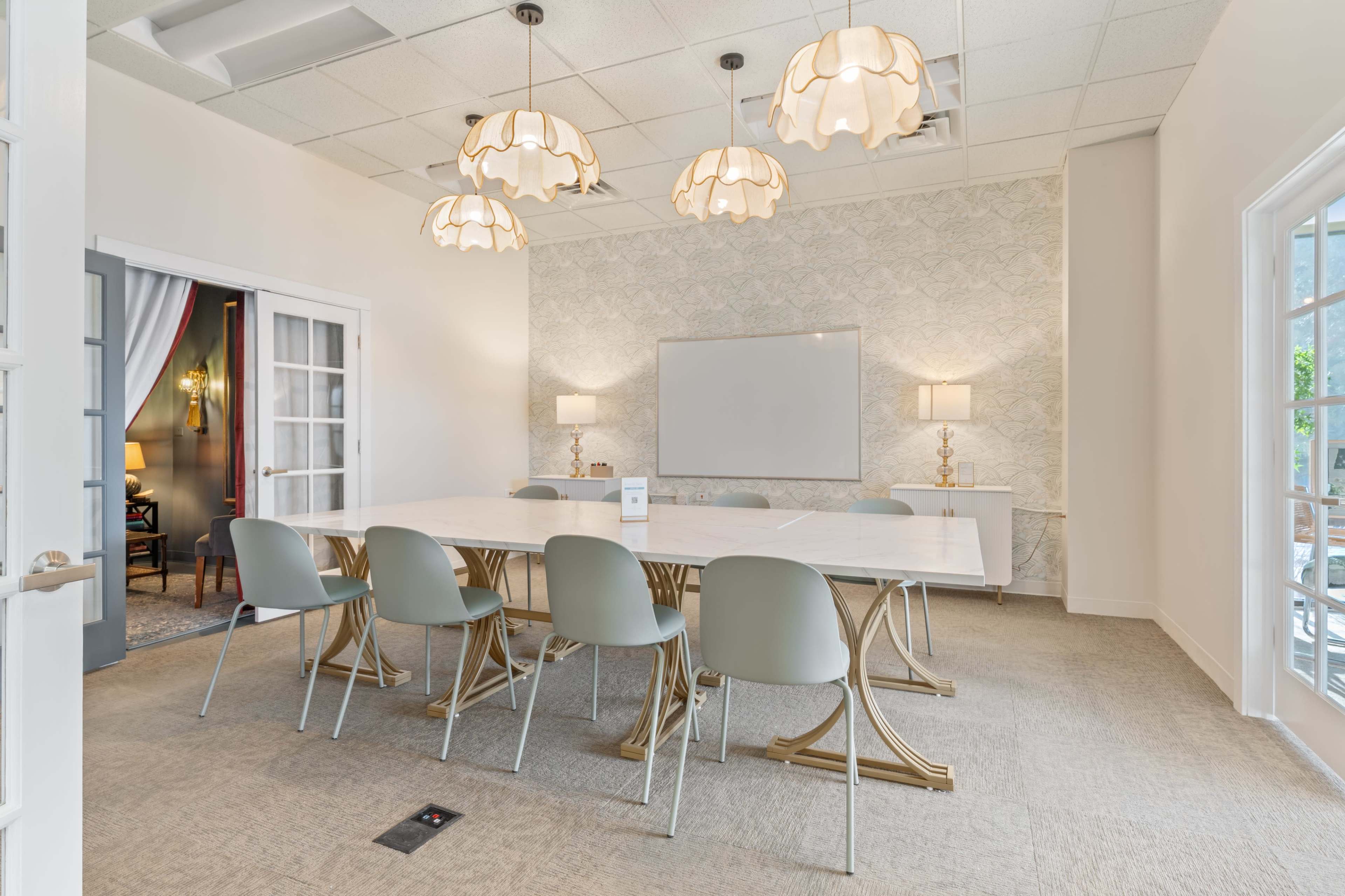 A conference room features a long table surrounded by gray chairs, with pendant lights overhead and a large whiteboard on the wall.