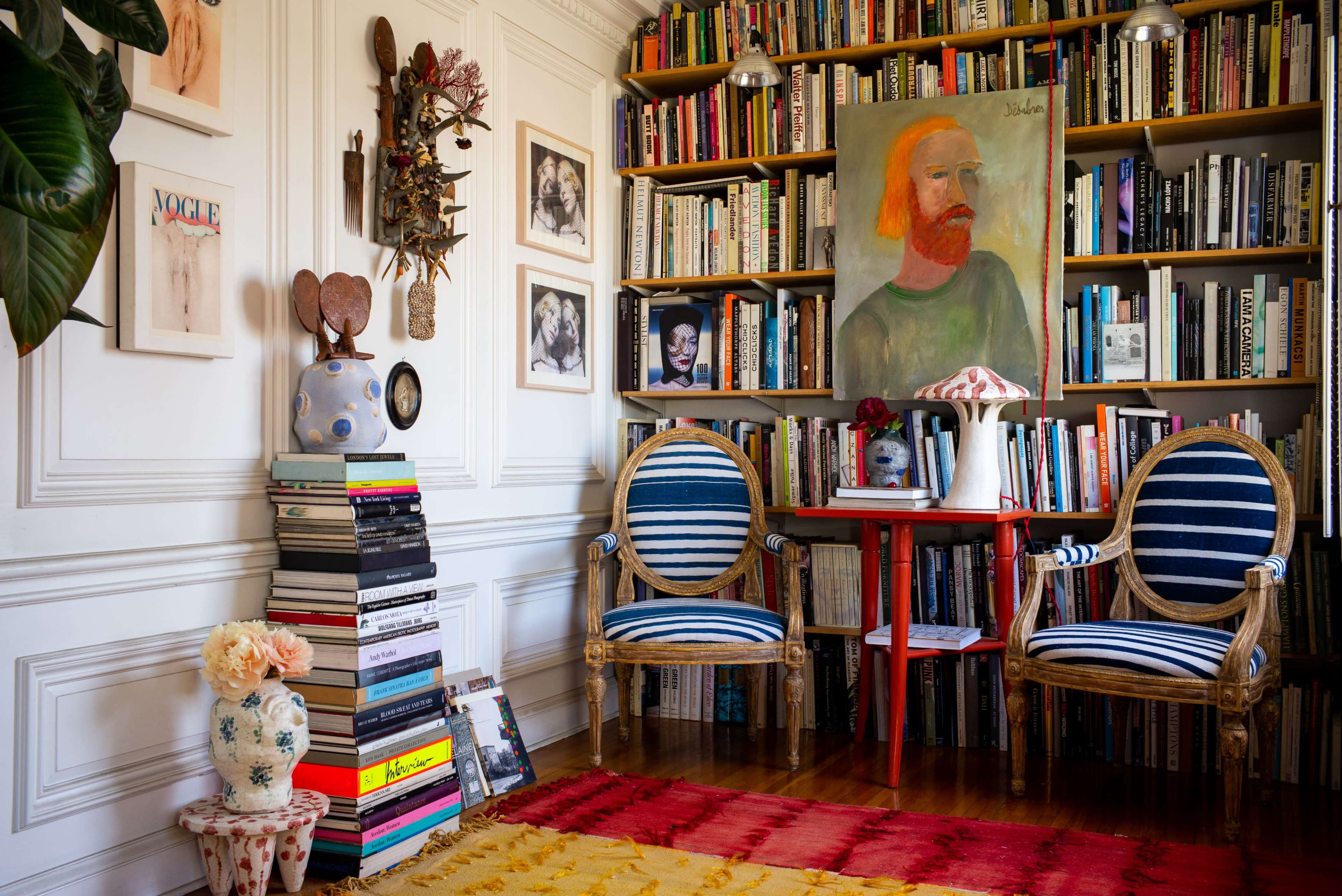 The image shows a cozy reading nook featuring two striped chairs, a small red table, a large bookshelf filled with books, and decorative elements on the walls.