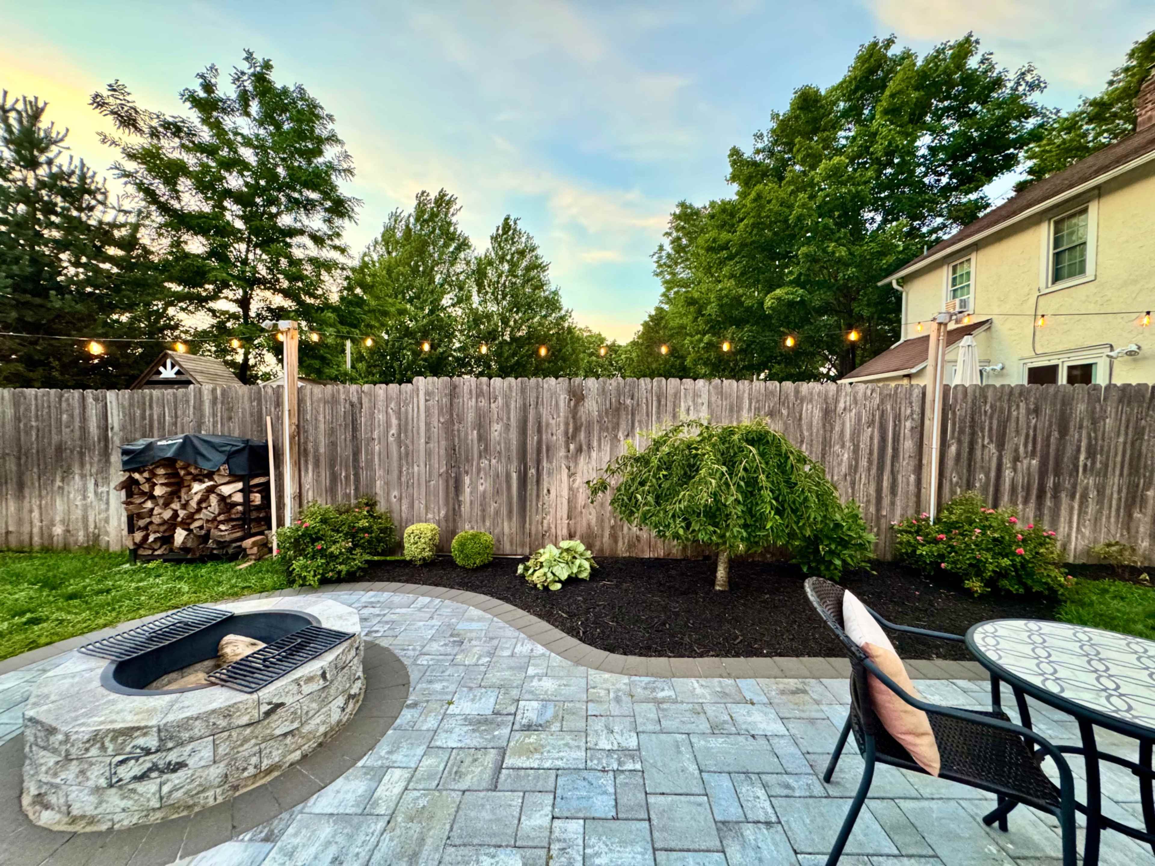The image depicts a backyard scene featuring a stone fire pit, a paved patio, landscaped grass, and a wooden fence adorned with string lights.