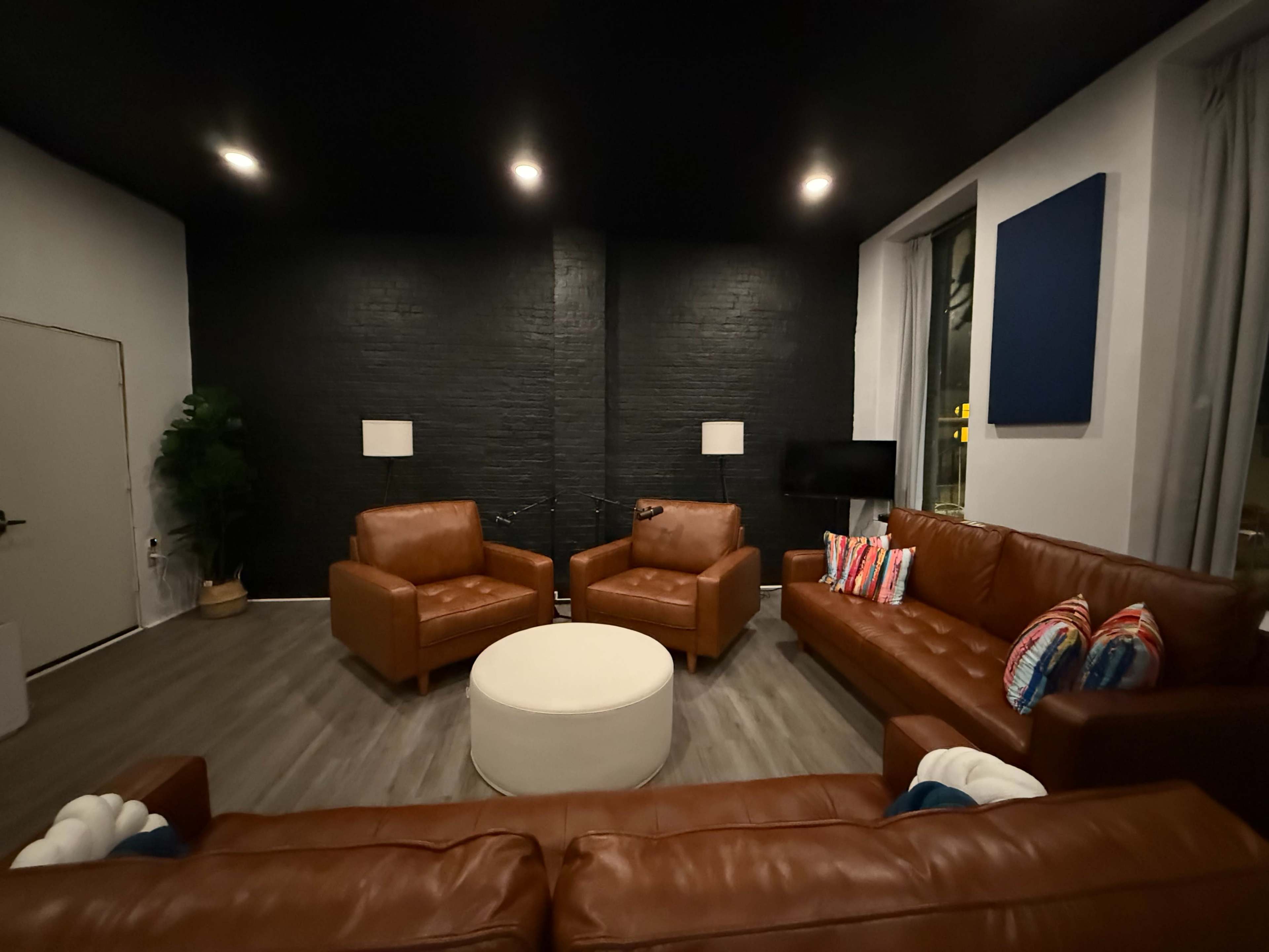 The image shows a modern lounge area with two brown leather armchairs, a brown leather sofa, a round white coffee table, and a television against a black brick wall.