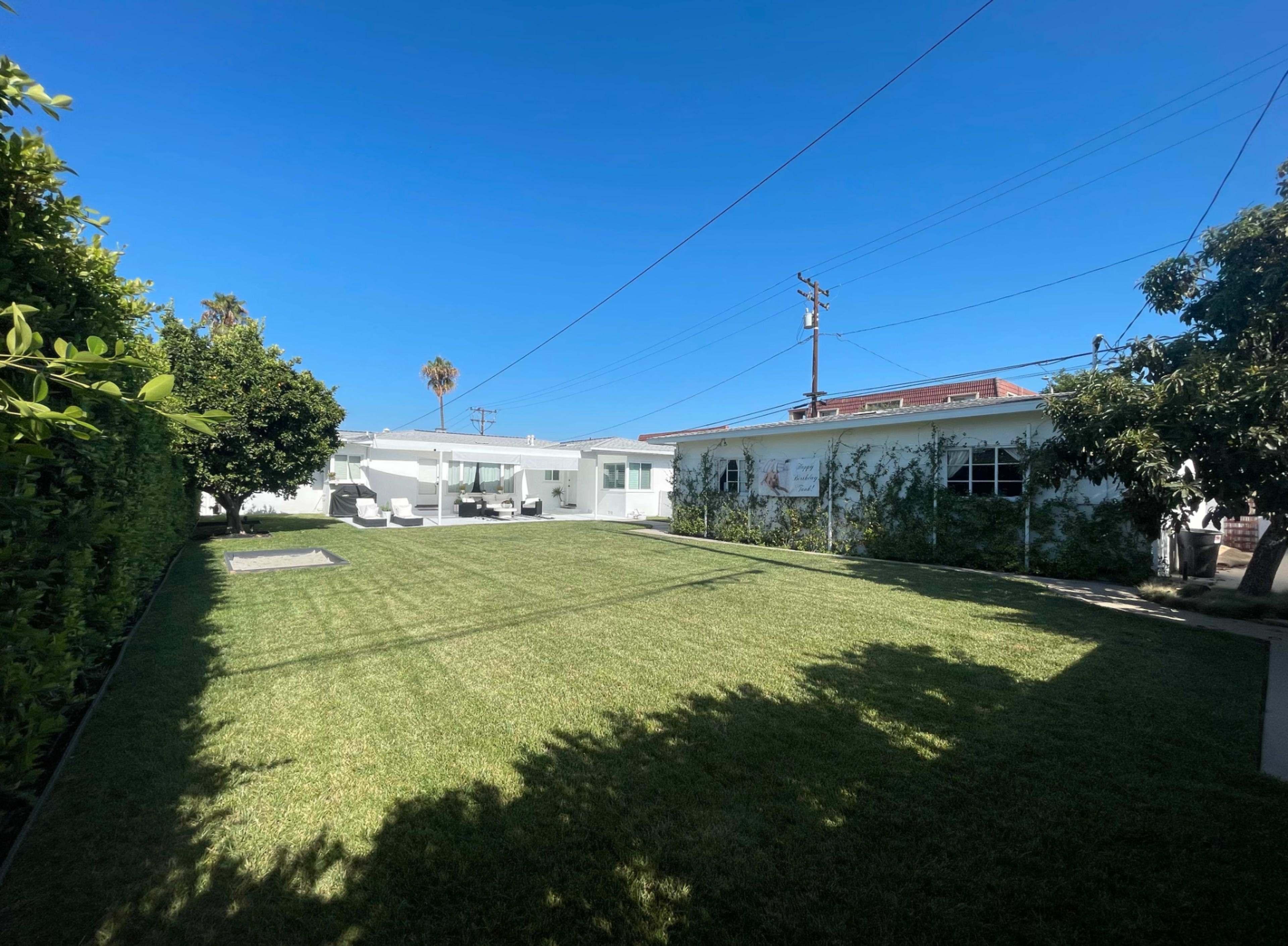 A well-maintained, green backyard with a clear blue sky, surrounded by trees and two white buildings in the background.