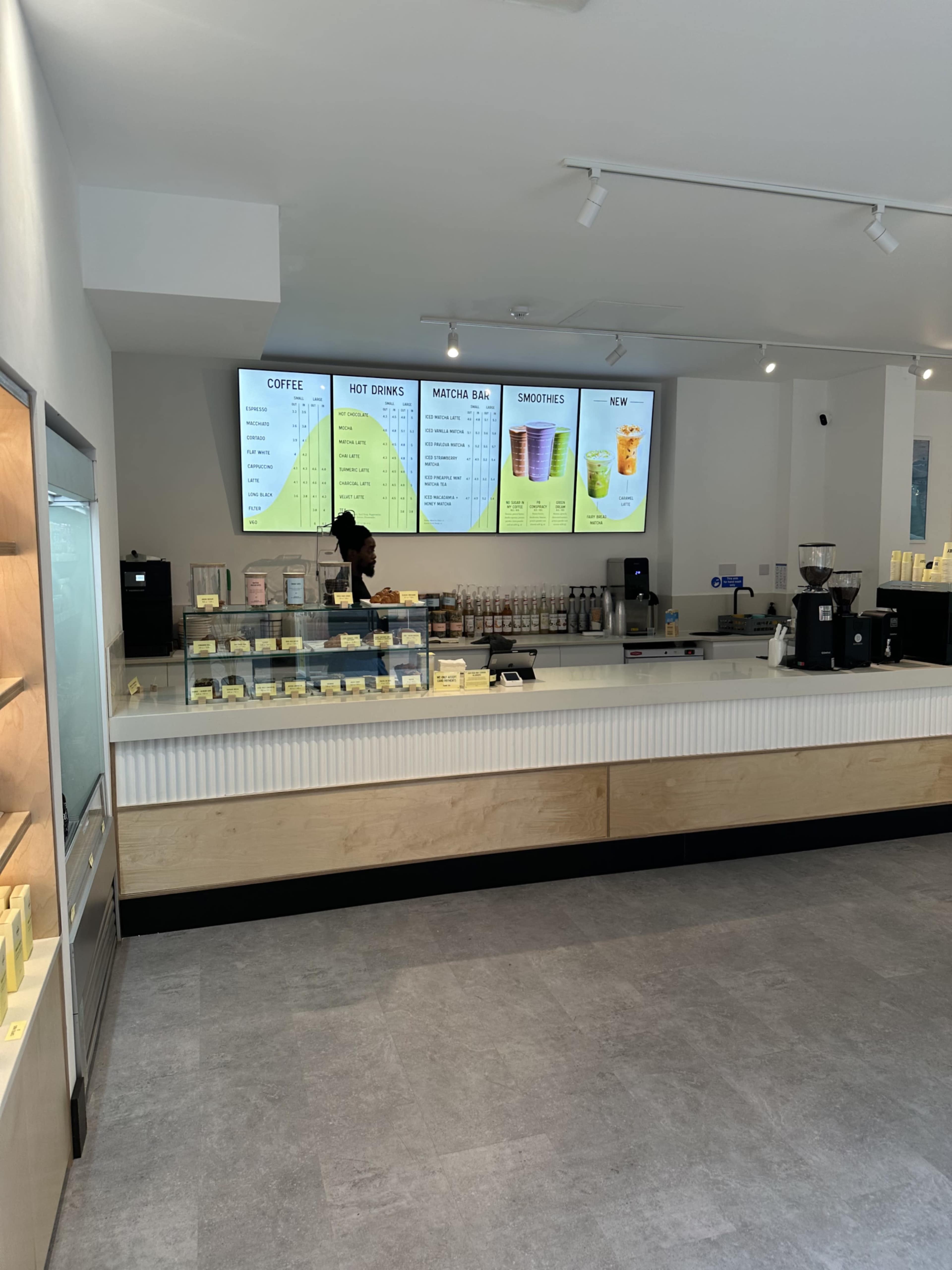 The image shows the interior of a modern café with a display counter, beverage menu boards, and various drink options visible on the shelves.