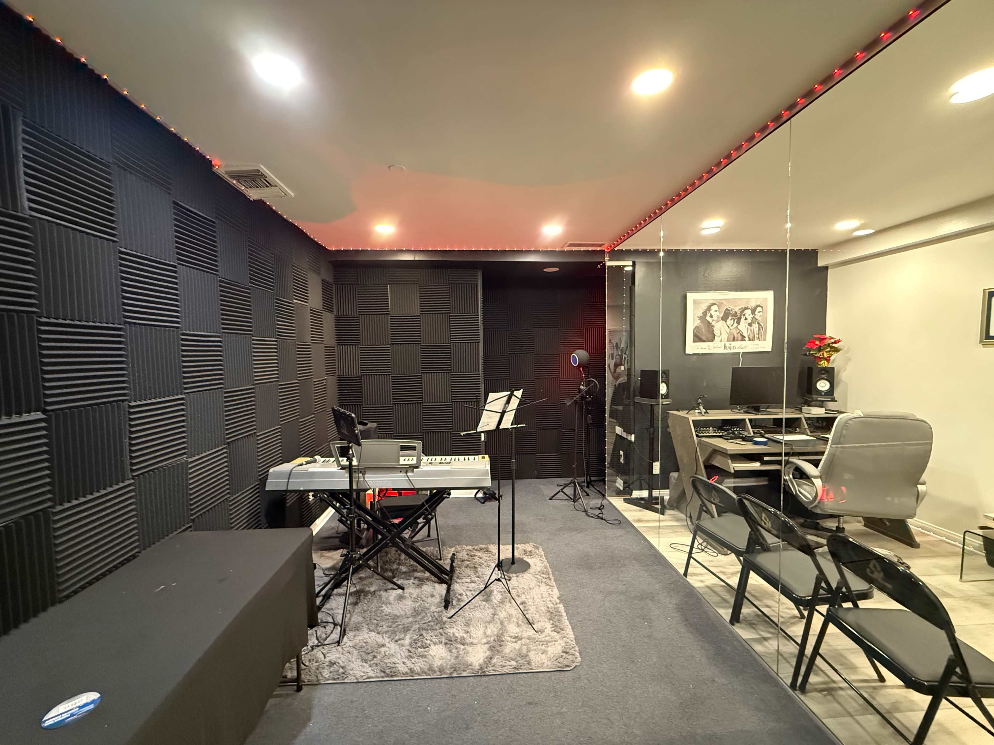 The image shows a music studio with soundproofed walls, a keyboard set up with a music stand, and a glass-enclosed workspace containing a computer and office chair.