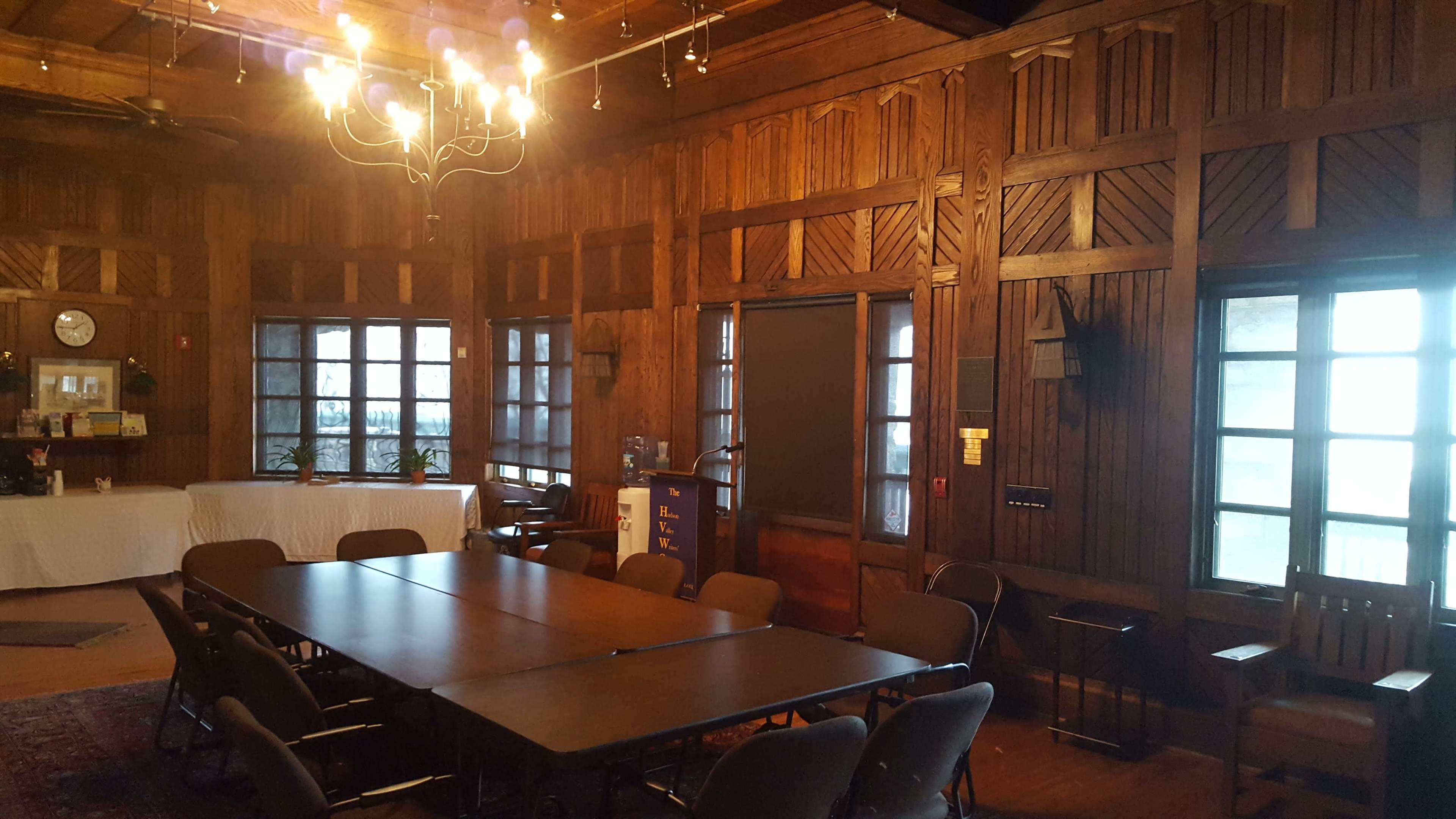 The image shows a wooden-paneled meeting room featuring a large table surrounded by chairs, with a chandelier overhead and windows providing light.
