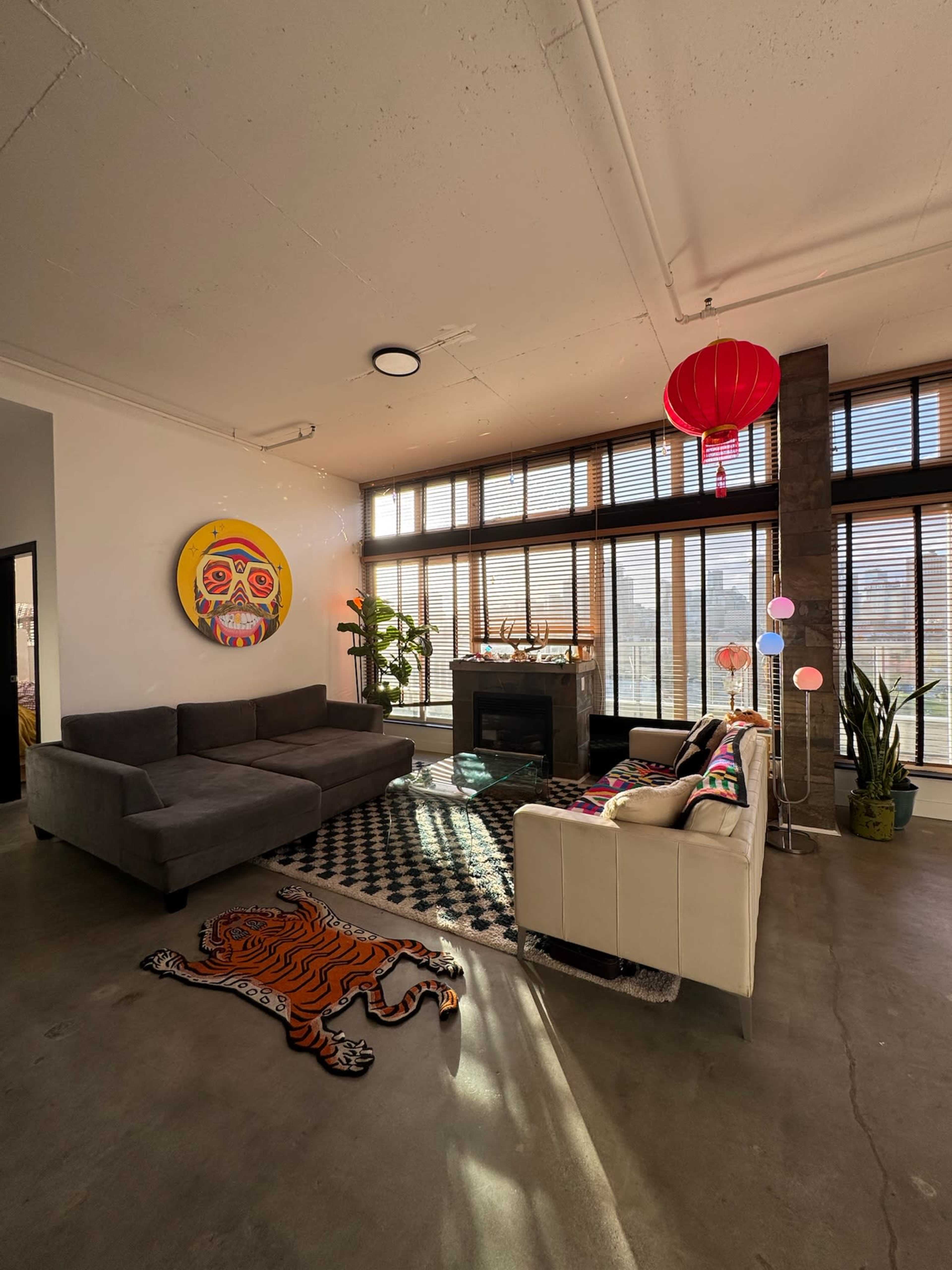 The image shows a modern living room with large windows, a gray sectional sofa, a white couch, and various decorative items, including a vibrant artwork on the wall and a tiger-shaped rug.