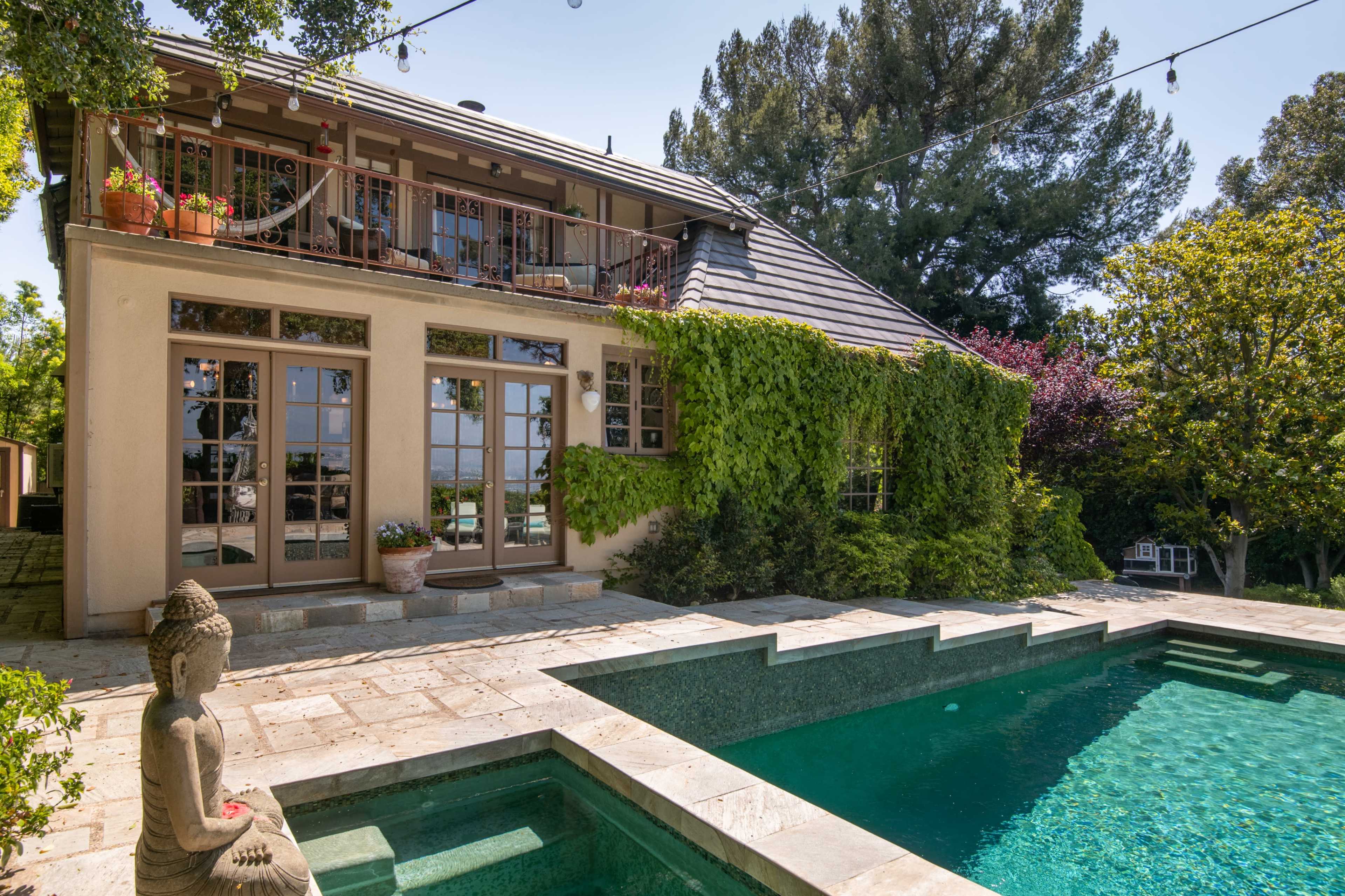 A two-story house with large windows, surrounded by greenery, next to a swimming pool with a Buddha statue at the edge.
