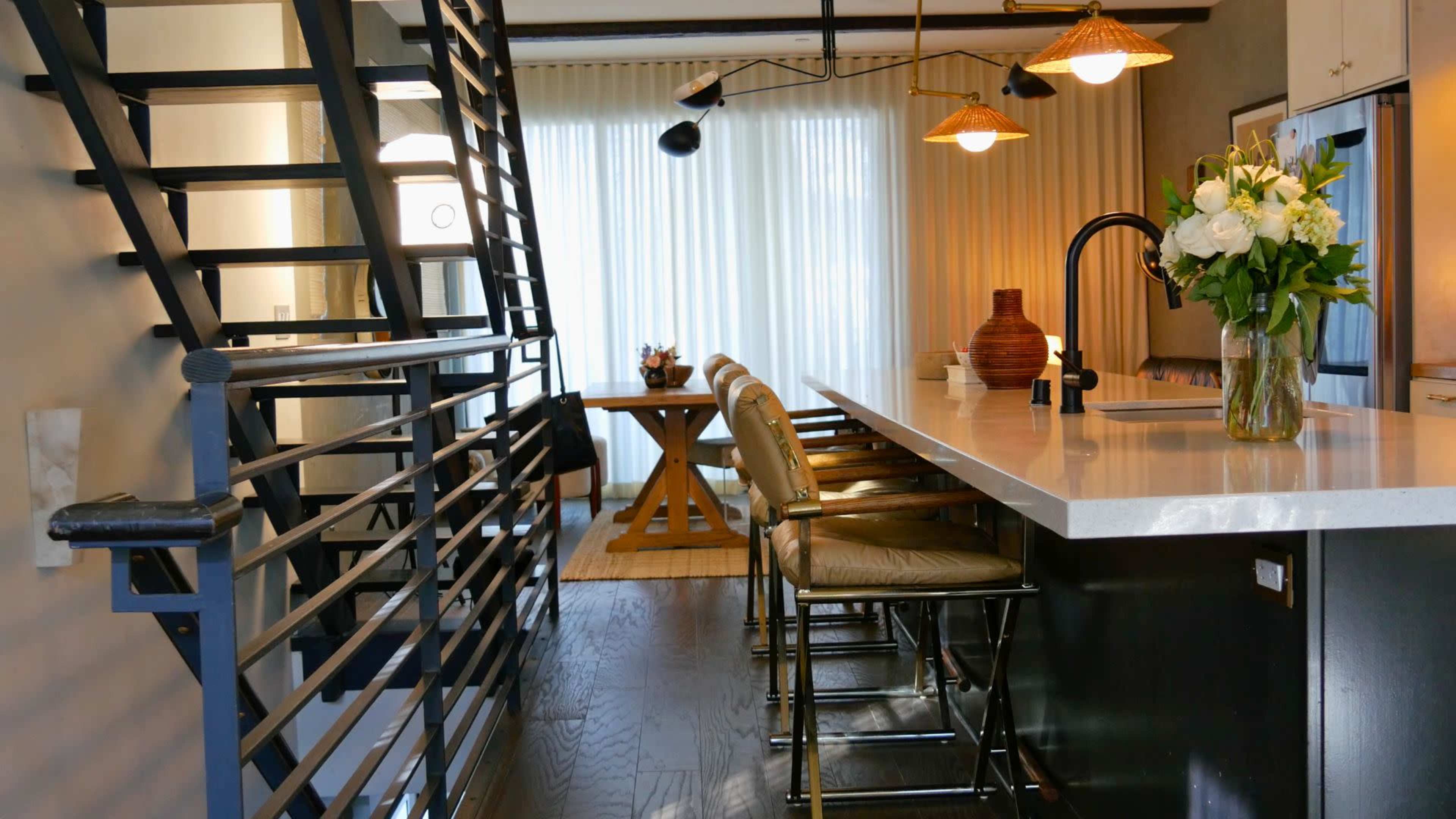The image shows a modern kitchen area with a staircase in the foreground, featuring bar stools at a countertop and large windows providing natural light.