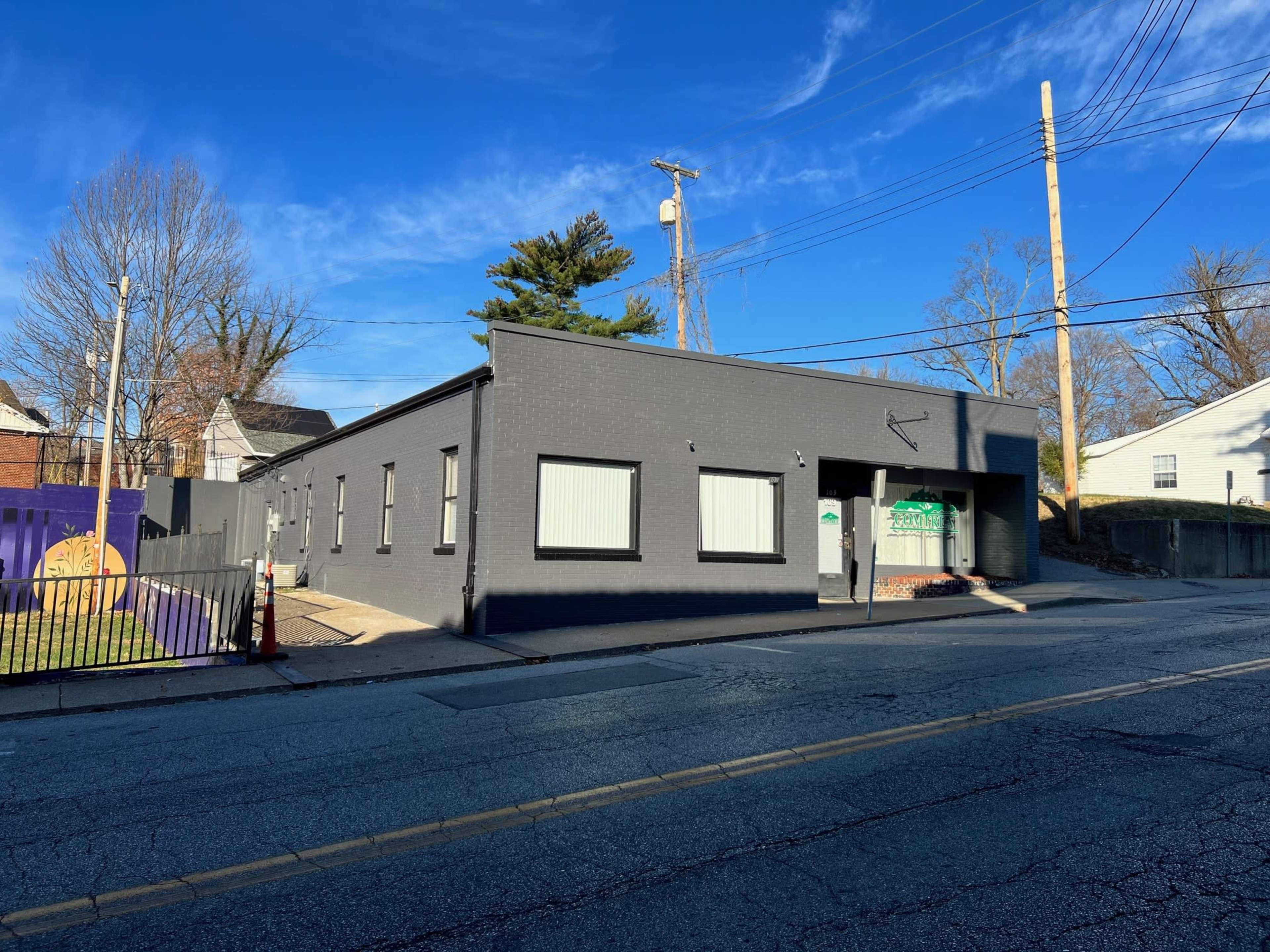 A gray building with large windows is situated on a street corner, with a fence and power lines visible in the background.