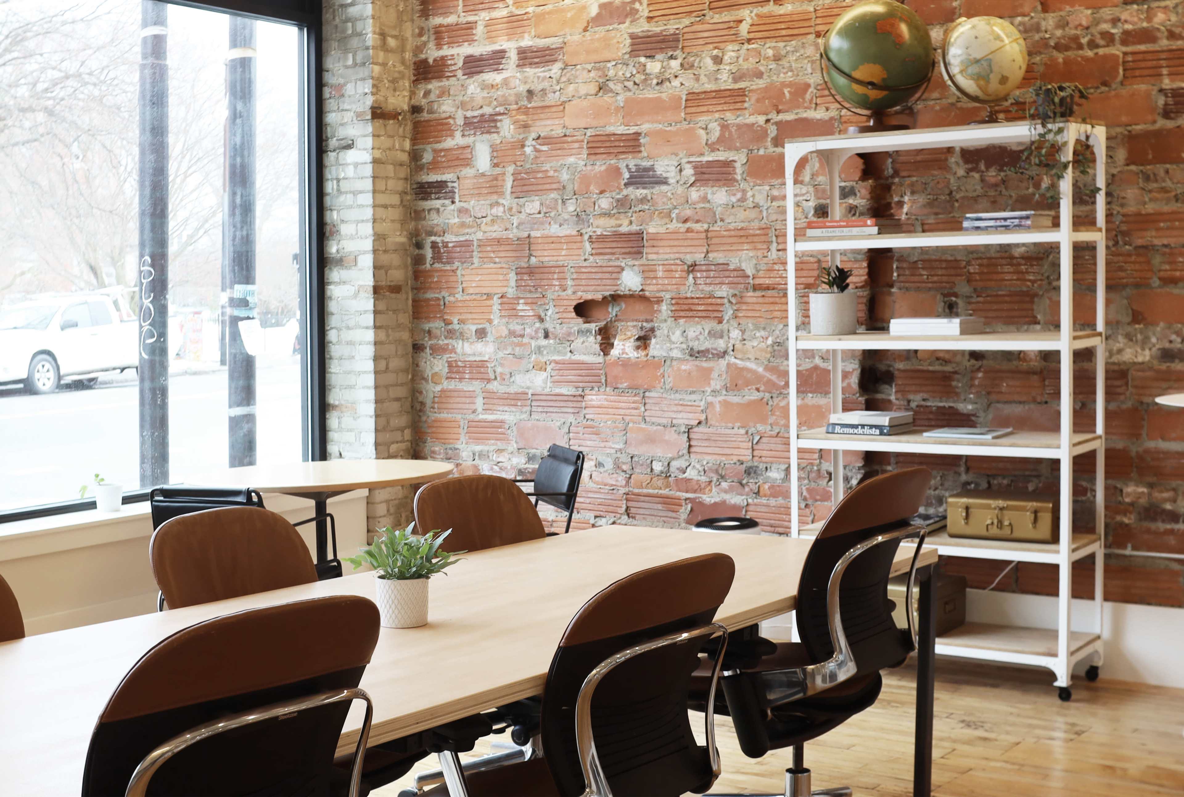 A modern meeting space with a wooden table surrounded by chairs, a brick wall in the background, and a shelf displaying plants and globes.