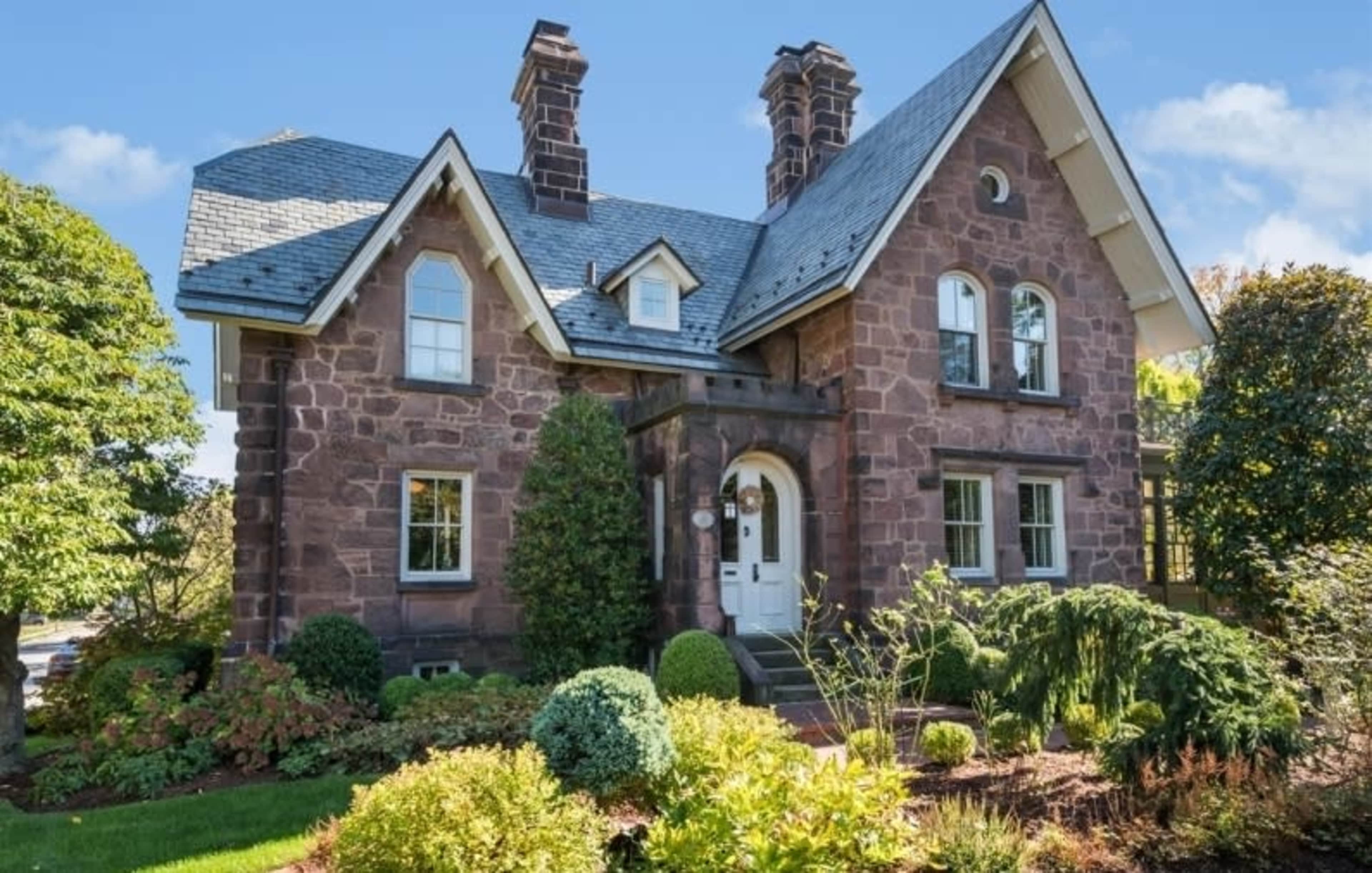 The image shows a stone house with a sloped roof and two chimneys, surrounded by well-maintained landscaping.