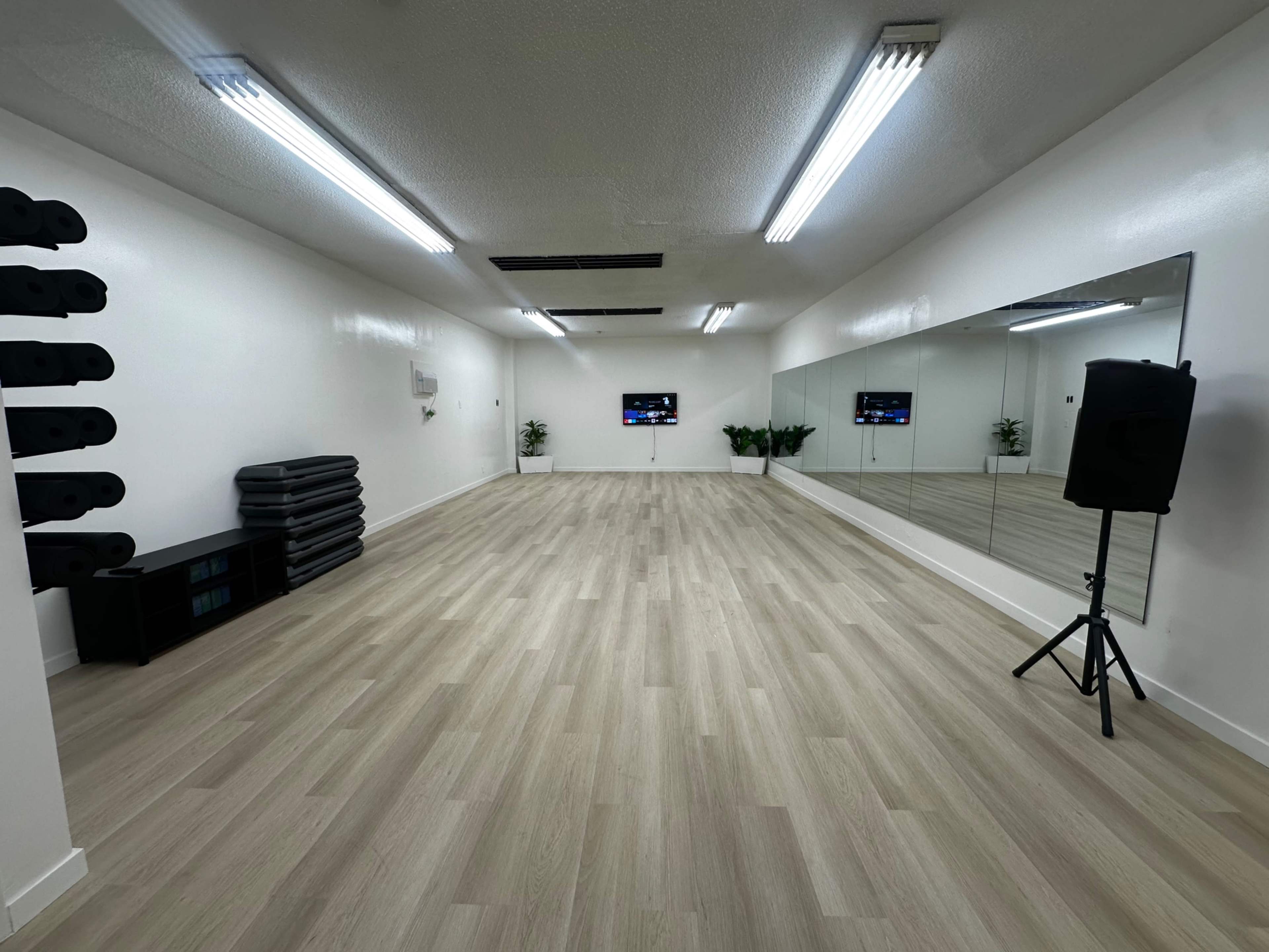 The image shows a spacious, empty room with wooden flooring, a mirrored wall, a small stage speaker, two television screens, and neatly stacked fitness equipment against one wall.