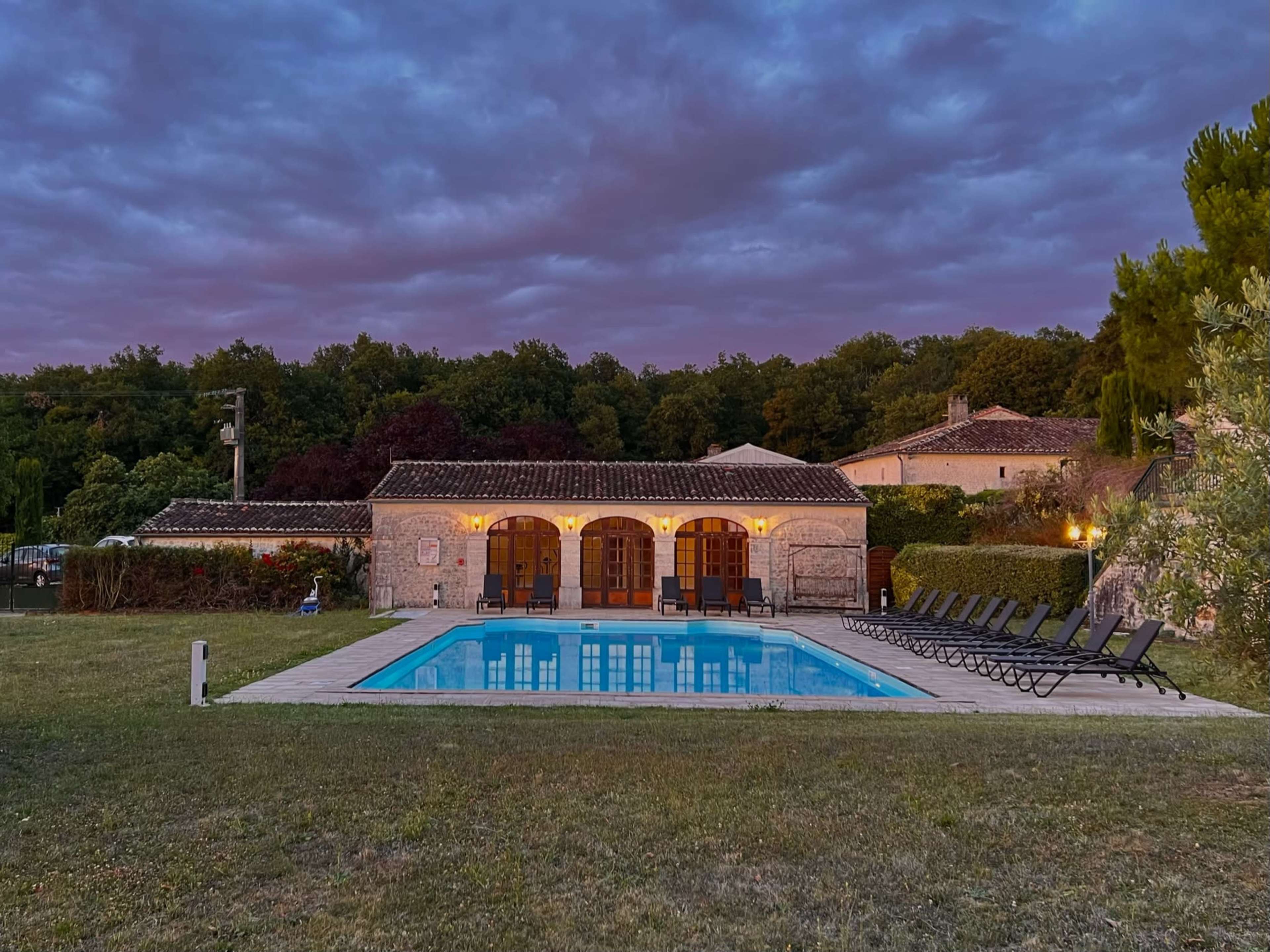 A stone villa with an outdoor swimming pool is set against a backdrop of darkening clouds and green trees.