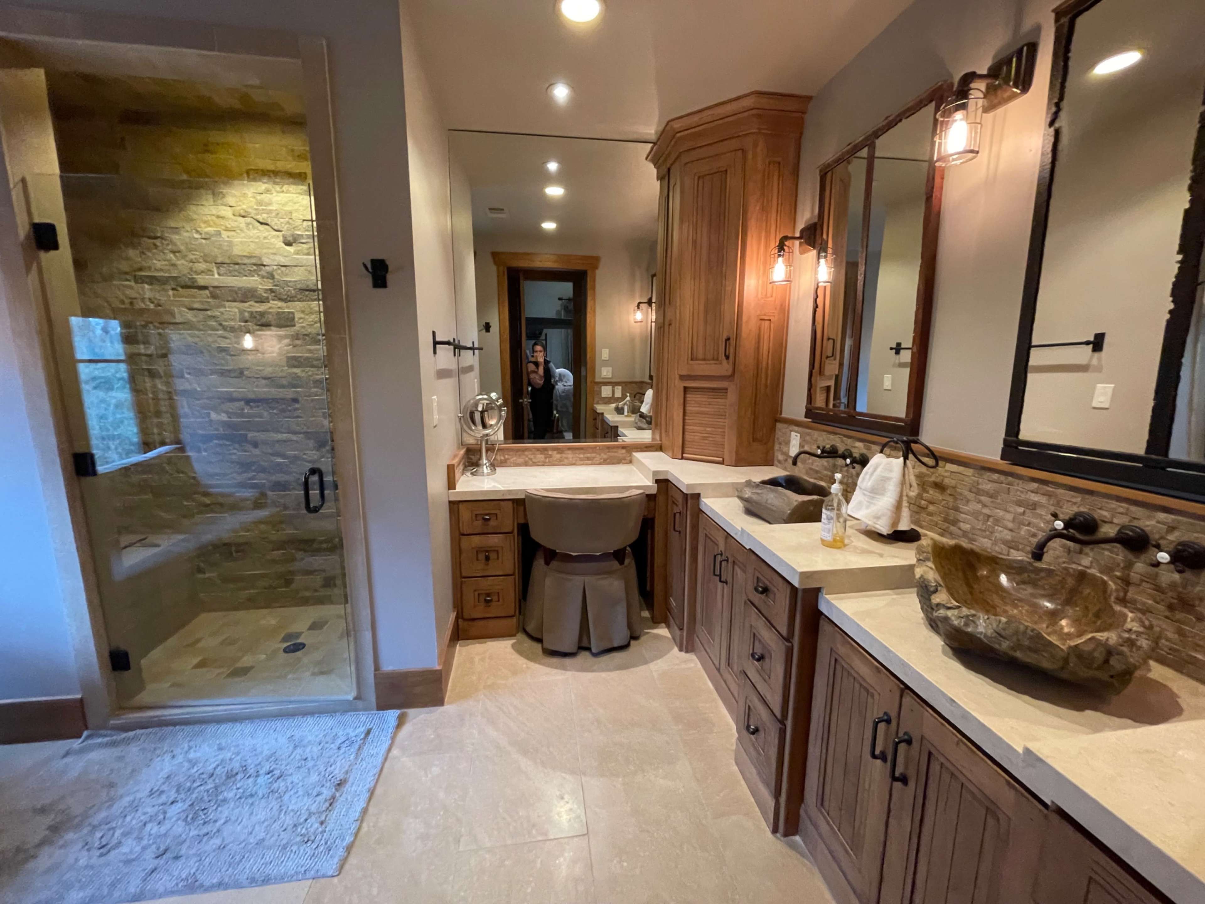 The image shows a spacious bathroom featuring a glass shower, a wooden vanity with a stone sink, and warm lighting.