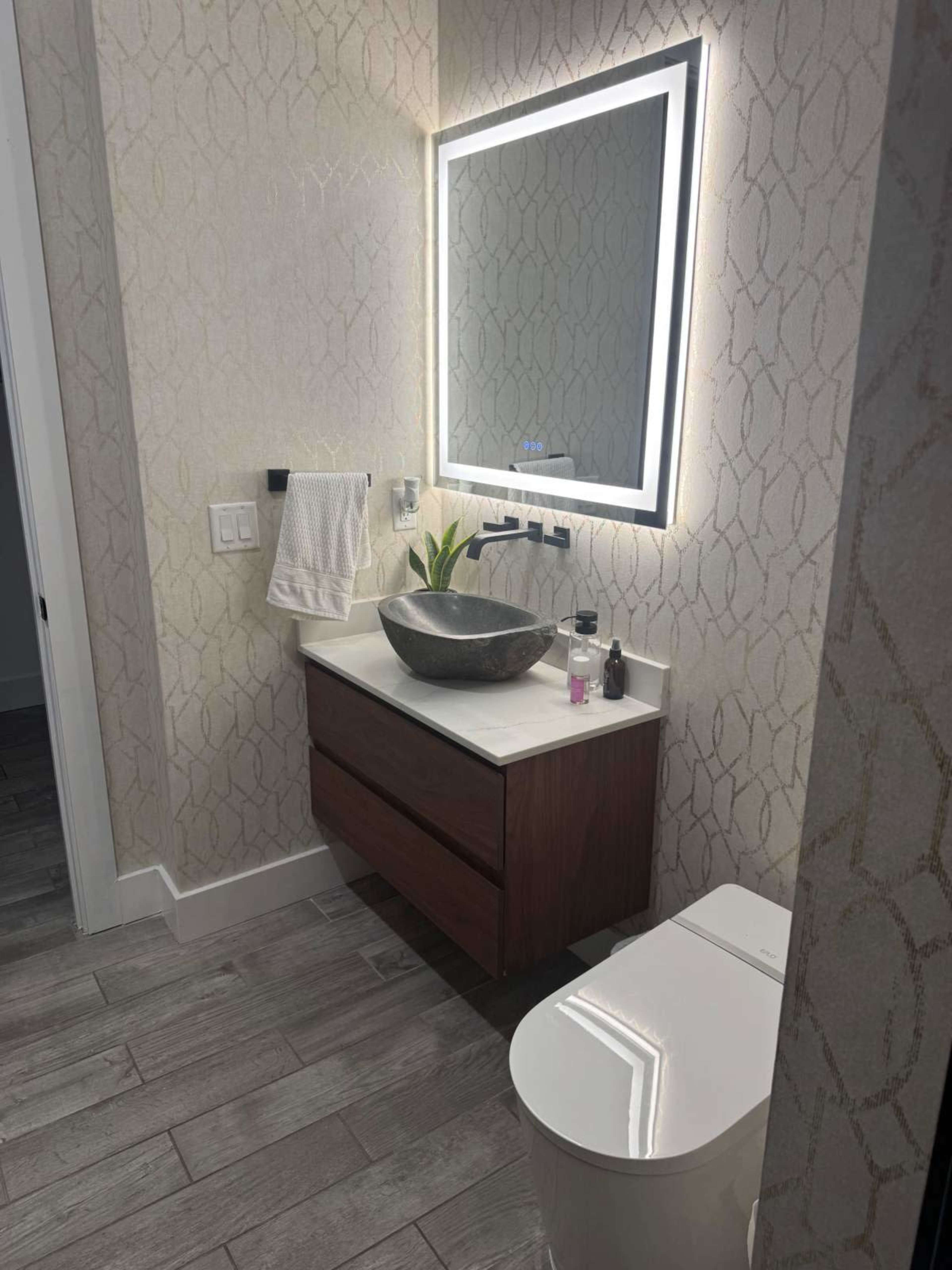 The image shows a modern bathroom featuring a lighted mirror above a stone sink with a wooden vanity and a white toilet.