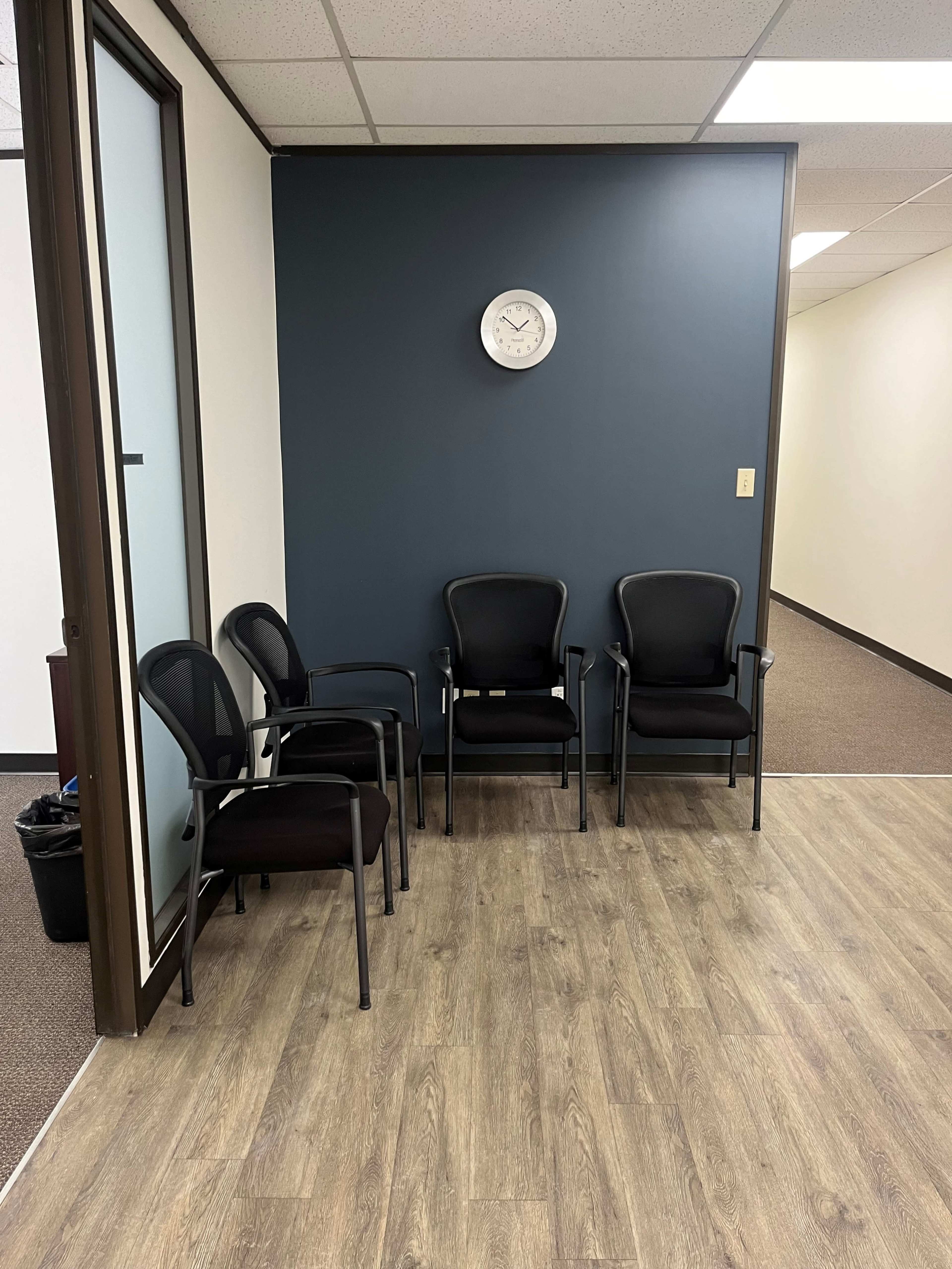 The image shows a waiting area featuring four black chairs against a dark blue wall, with a clock and a doorway leading to another room.
