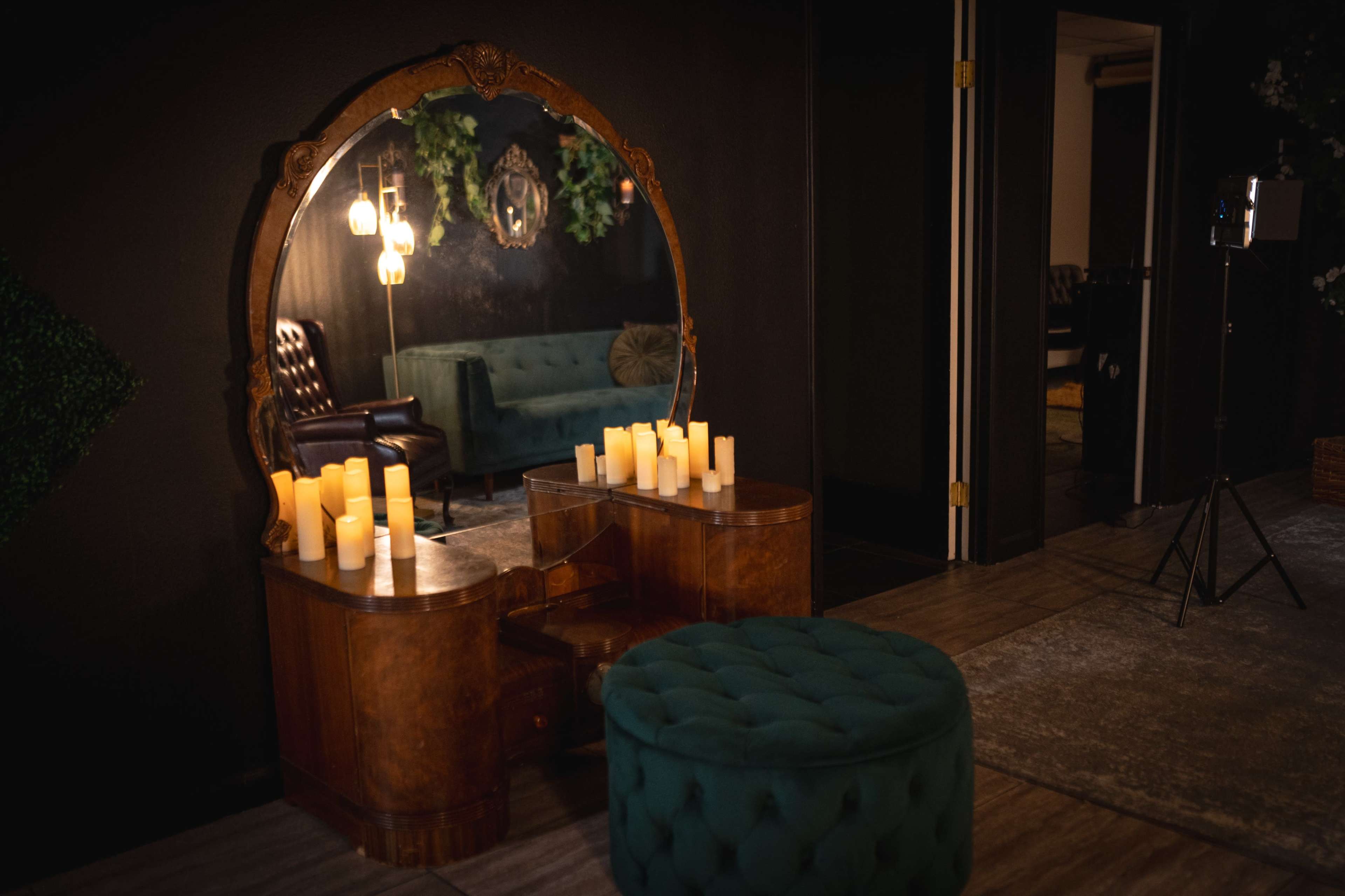 A vintage-style vanity table with candles sits in front of a large round mirror, alongside a green velvet ottoman and a dark-colored sofa in a dimly lit room.
