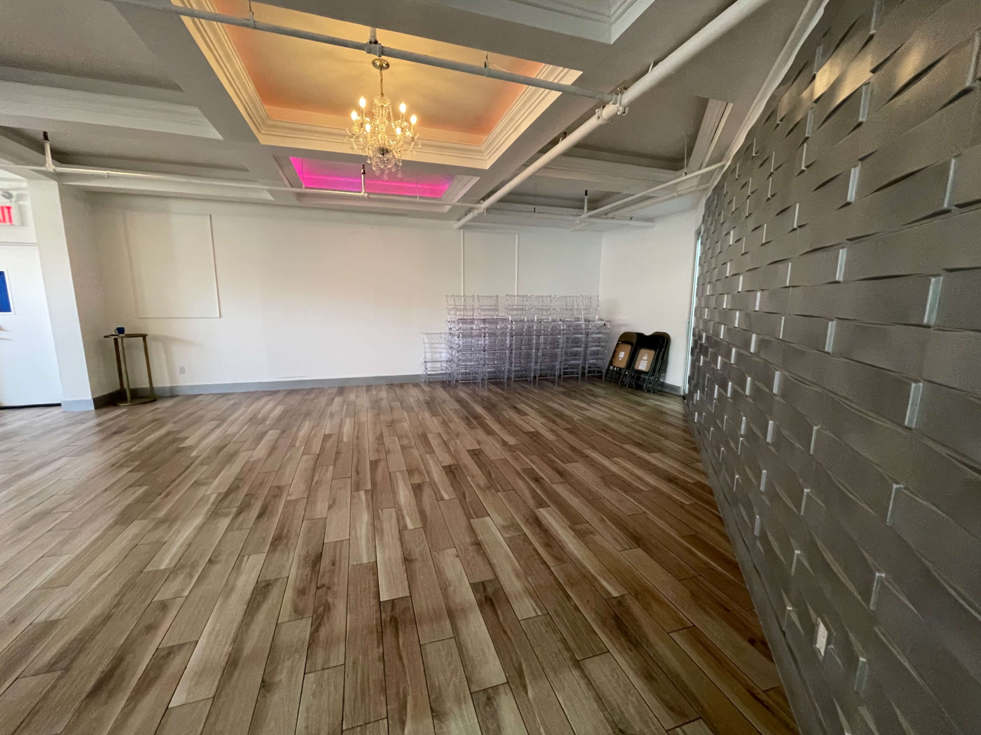 A spacious, empty room with wooden flooring and a wall featuring a patterned design, illuminated by a chandelier and soft lighting.