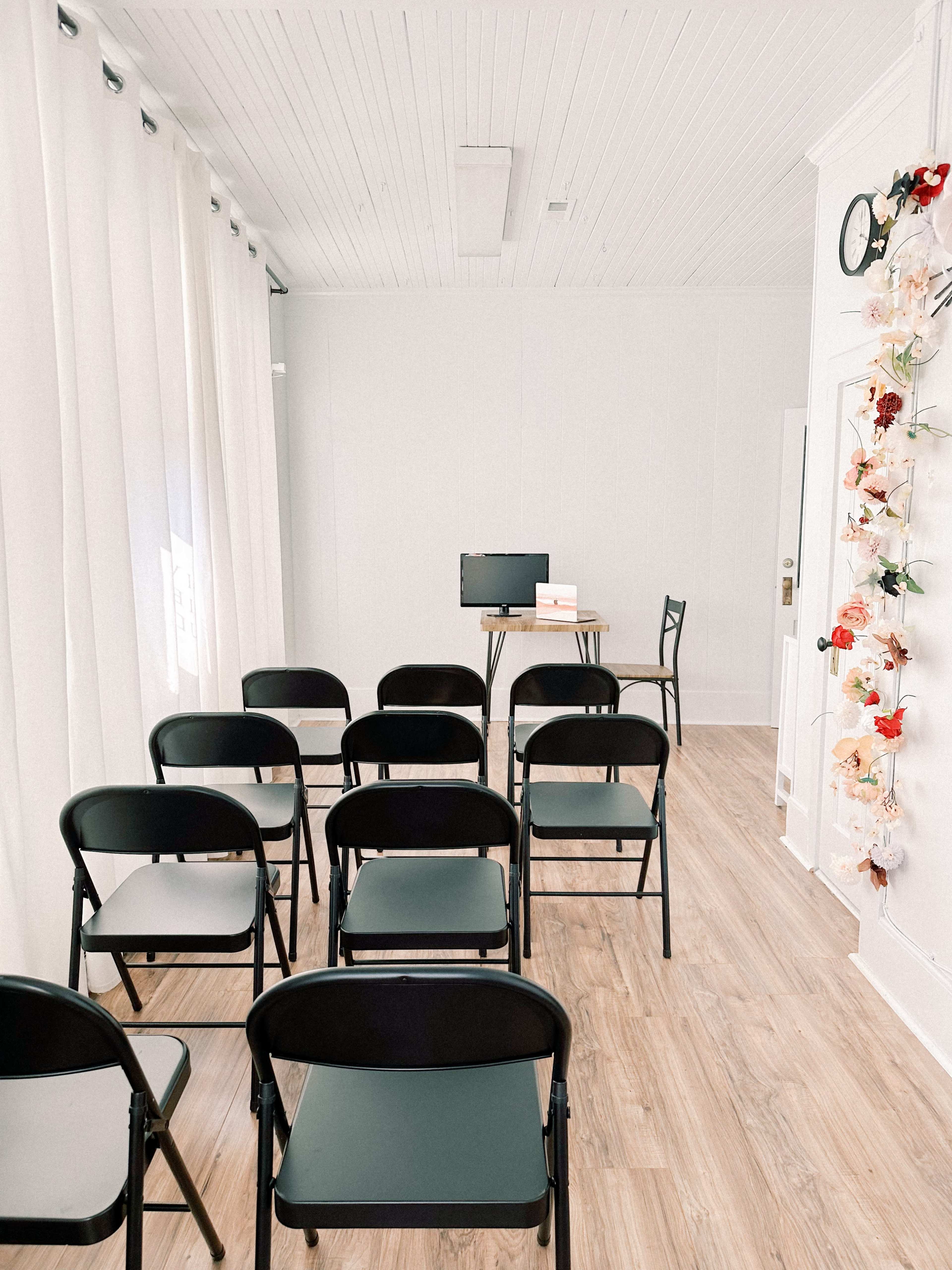 A room with folding black chairs arranged in rows facing a small table and a computer against a white wall, featuring decorative flowers and curtains.