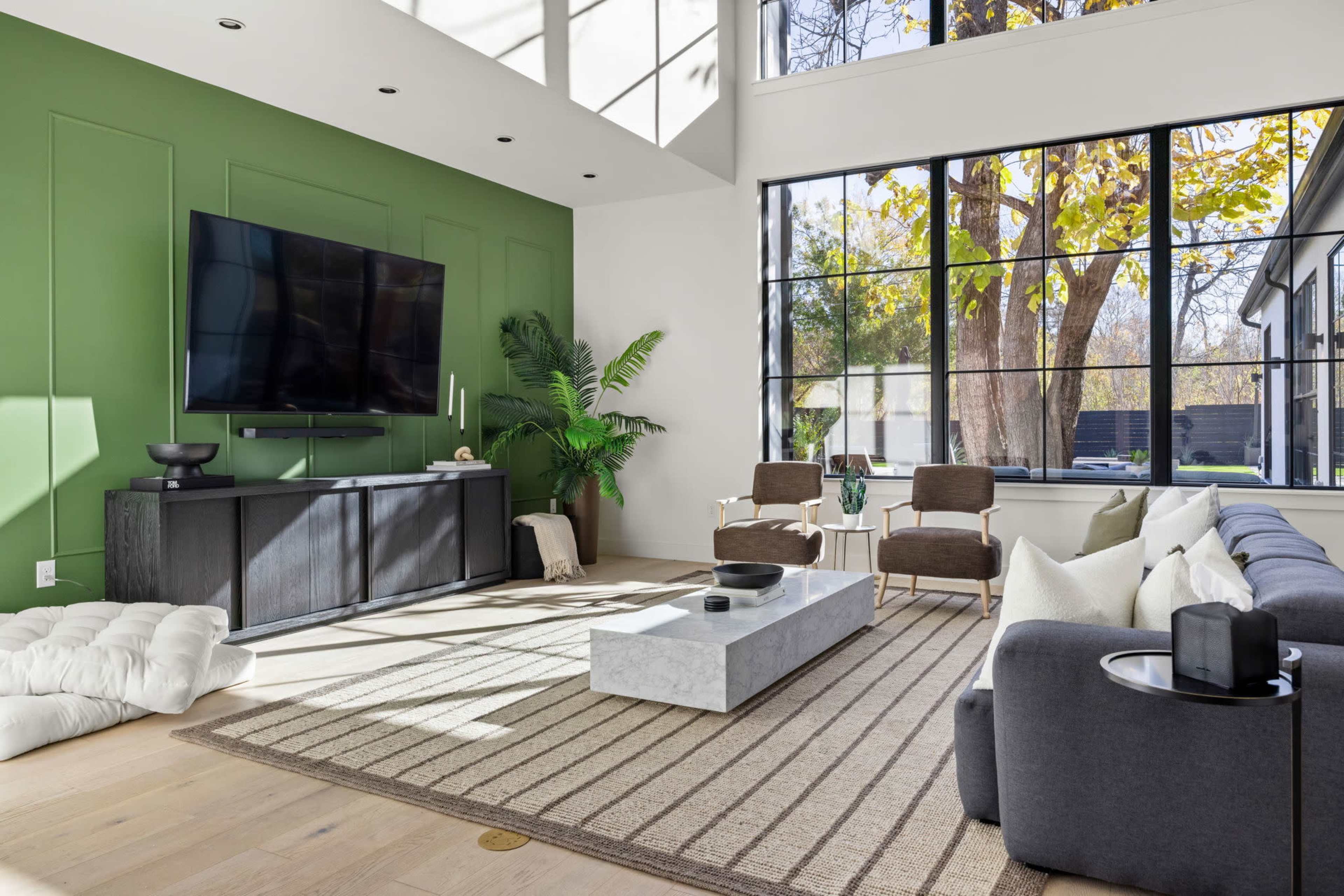 The room features a spacious living area with a large television mounted on a green accent wall, a gray sectional couch, two armchairs, and a decorative coffee table.