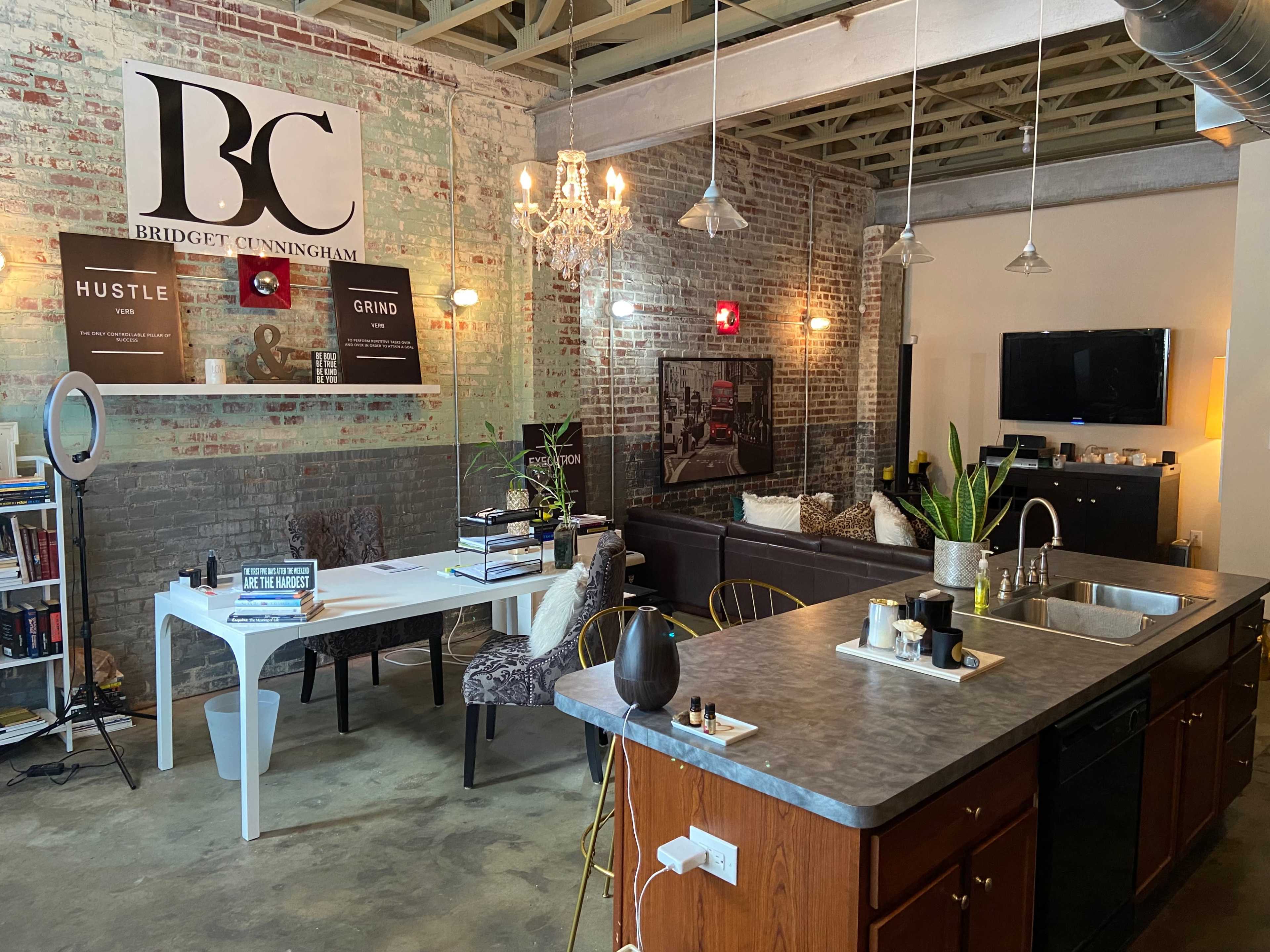 A modern interior space featuring a kitchen island with bar stools, a dining area with a white table, and a lounge area with a sofa, all set against exposed brick walls.
