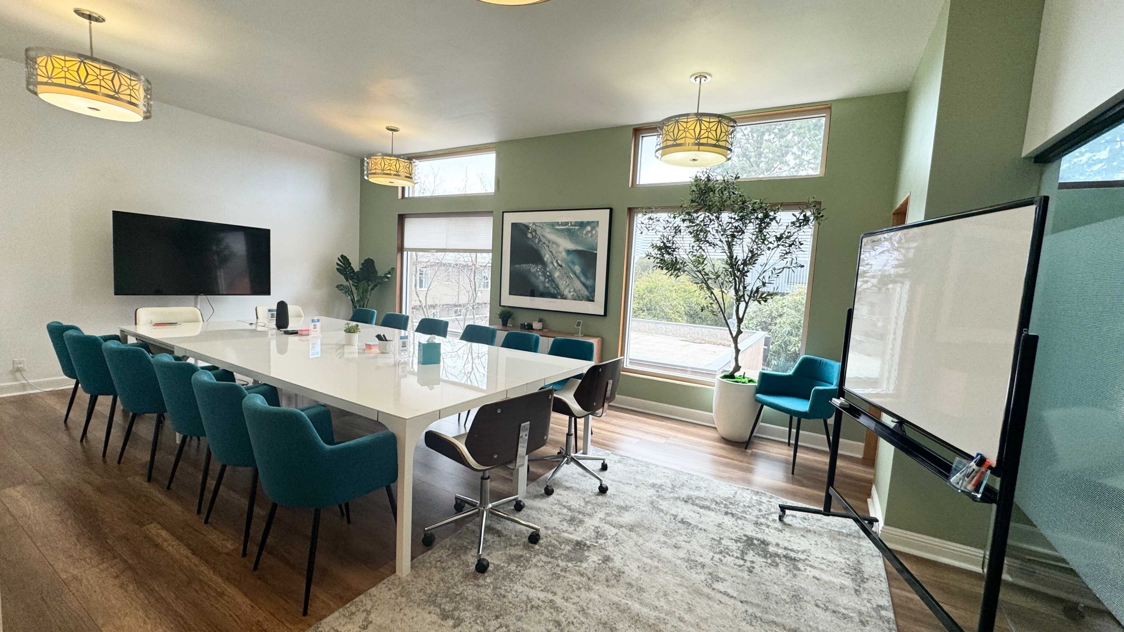 A modern conference room features a long white table surrounded by teal chairs, with large windows letting in natural light and greenery visible outside.