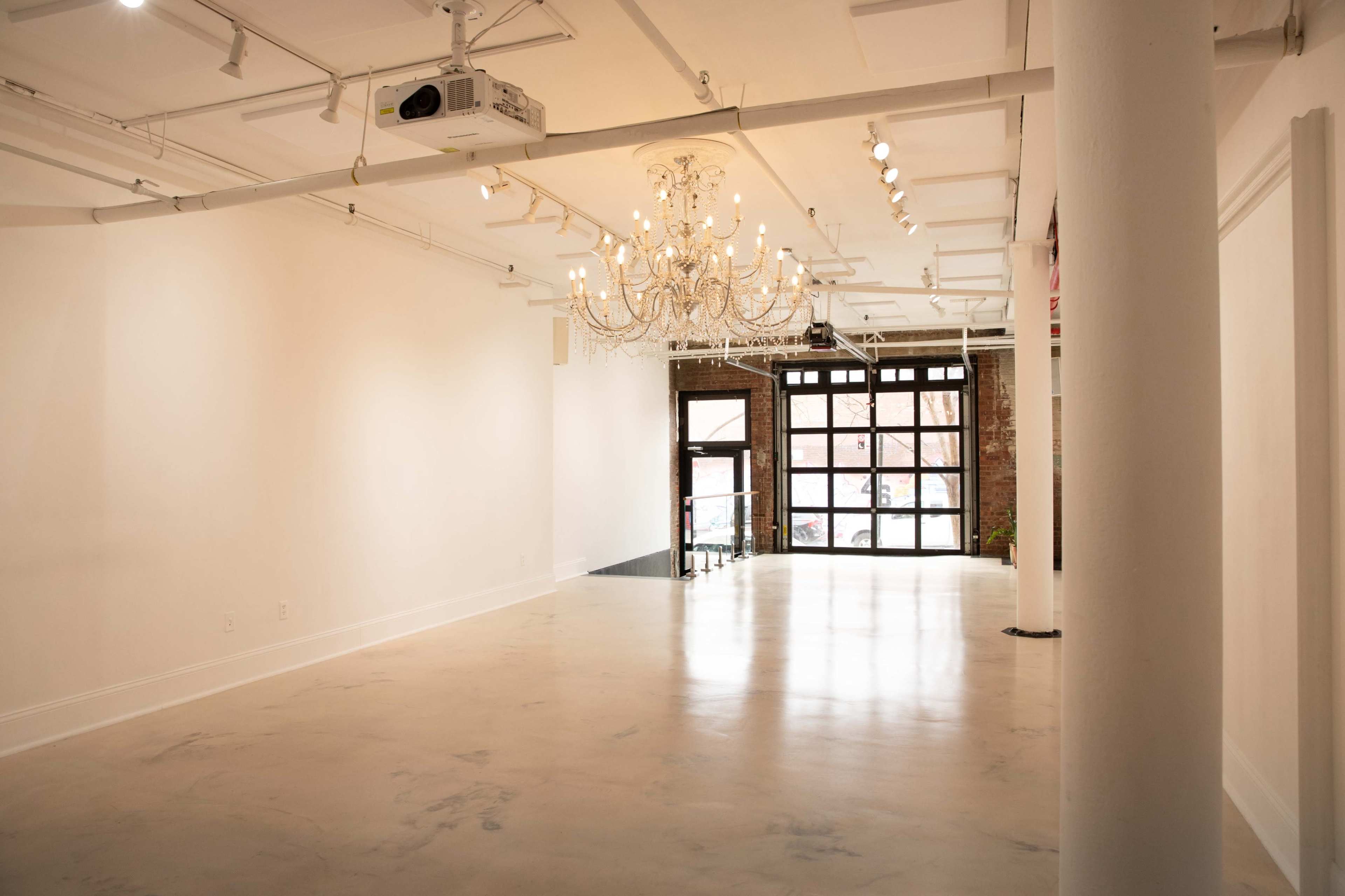 The image shows a spacious, empty room with a chandelier and large windows facing a street.