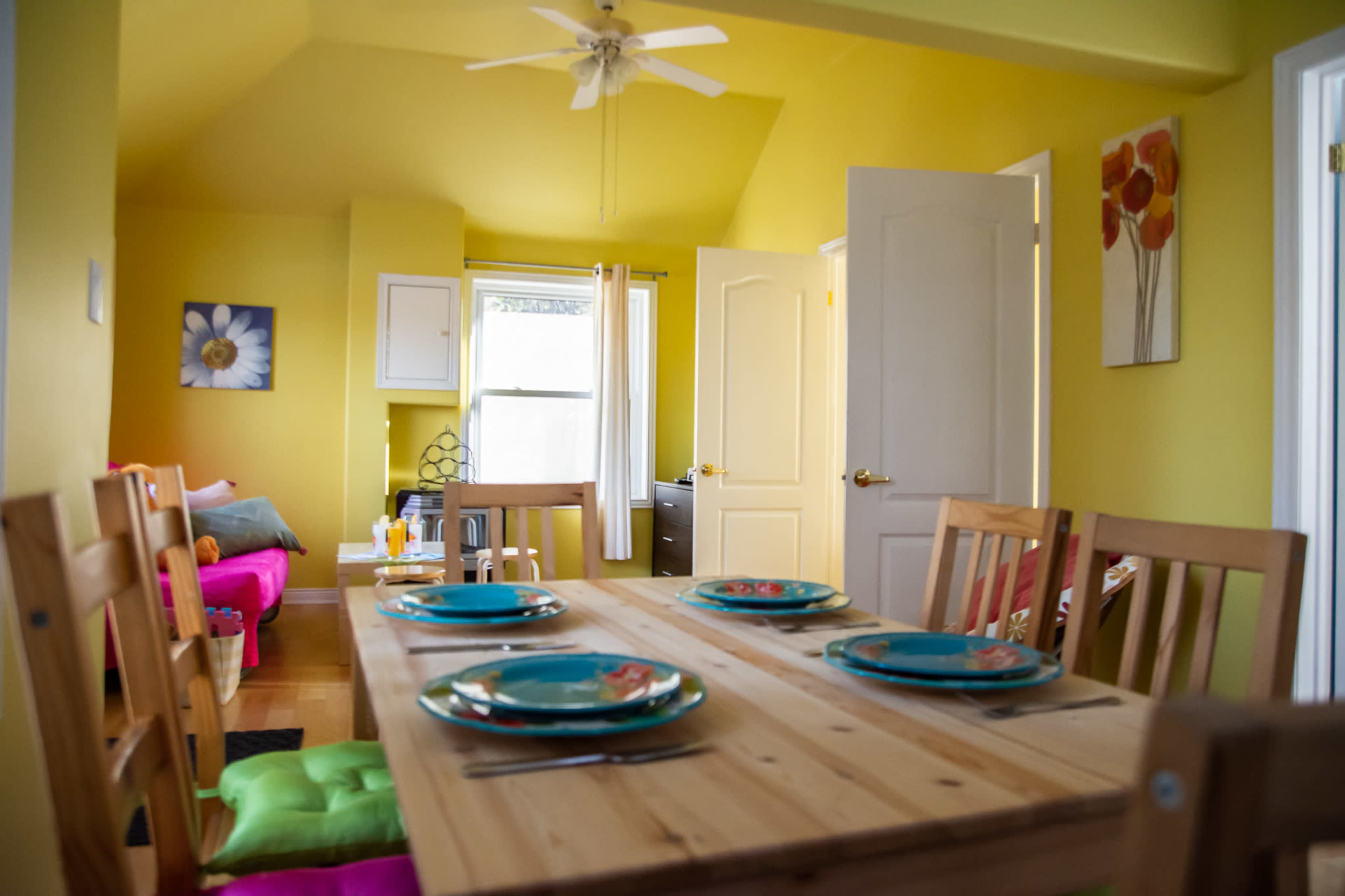 A bright yellow room features a wooden dining table set with decorative plates, chairs, and a view of a kitchen and living area beyond.