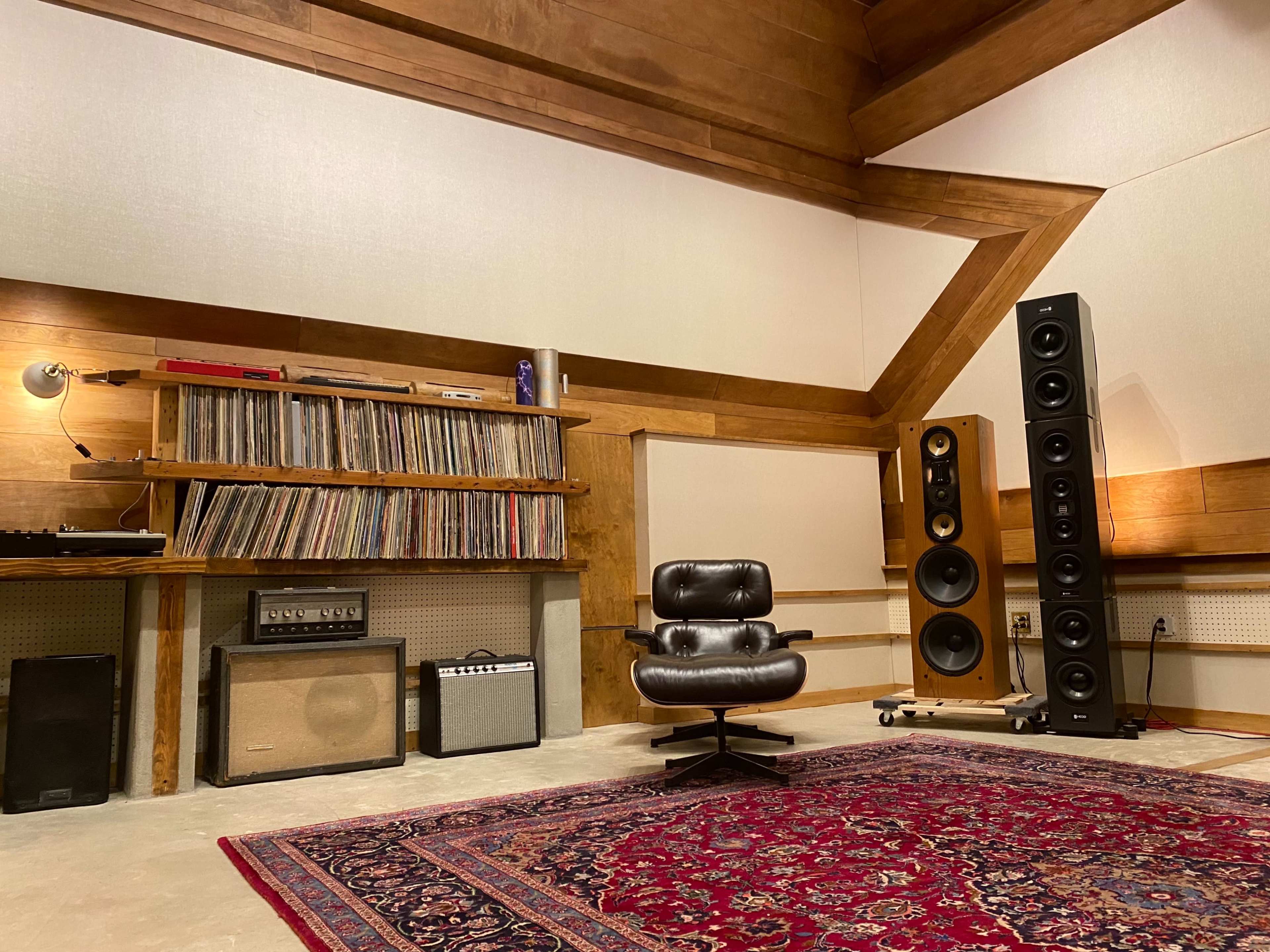 The image shows a music studio with a black swivel chair, shelves filled with vinyl records, a vintage amplifier, and tall speakers against a wooden wall.