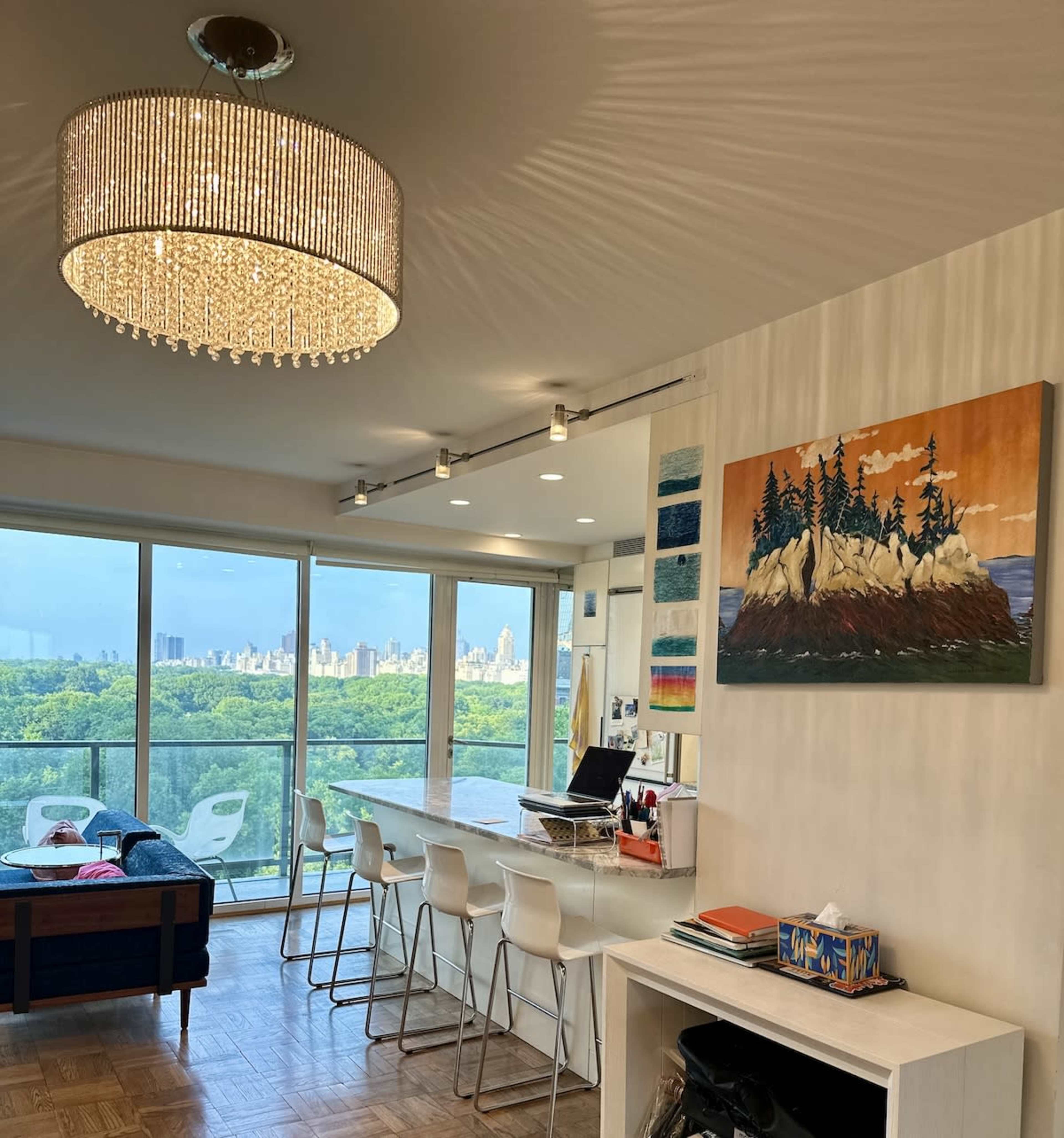 A modern interior features a chandelier, bar seating, and a large window offering a view of a city skyline and greenery.