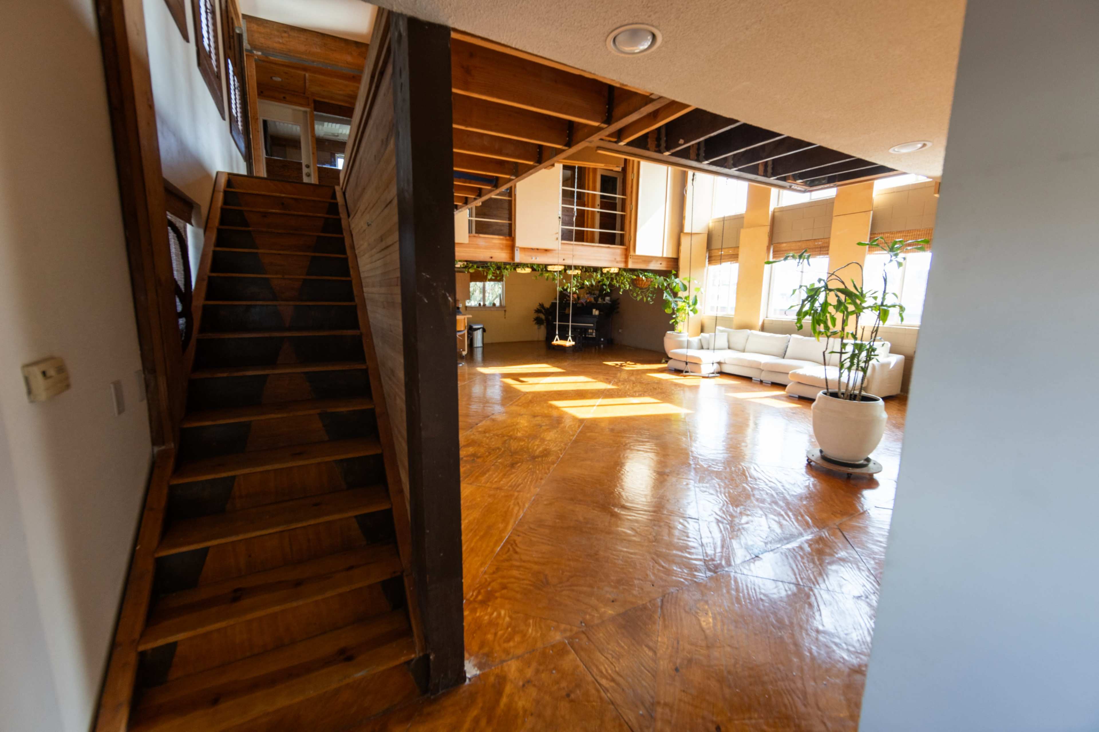 The image shows an interior space featuring a staircase leading to a second level and a bright living area furnished with a sofa and greenery.