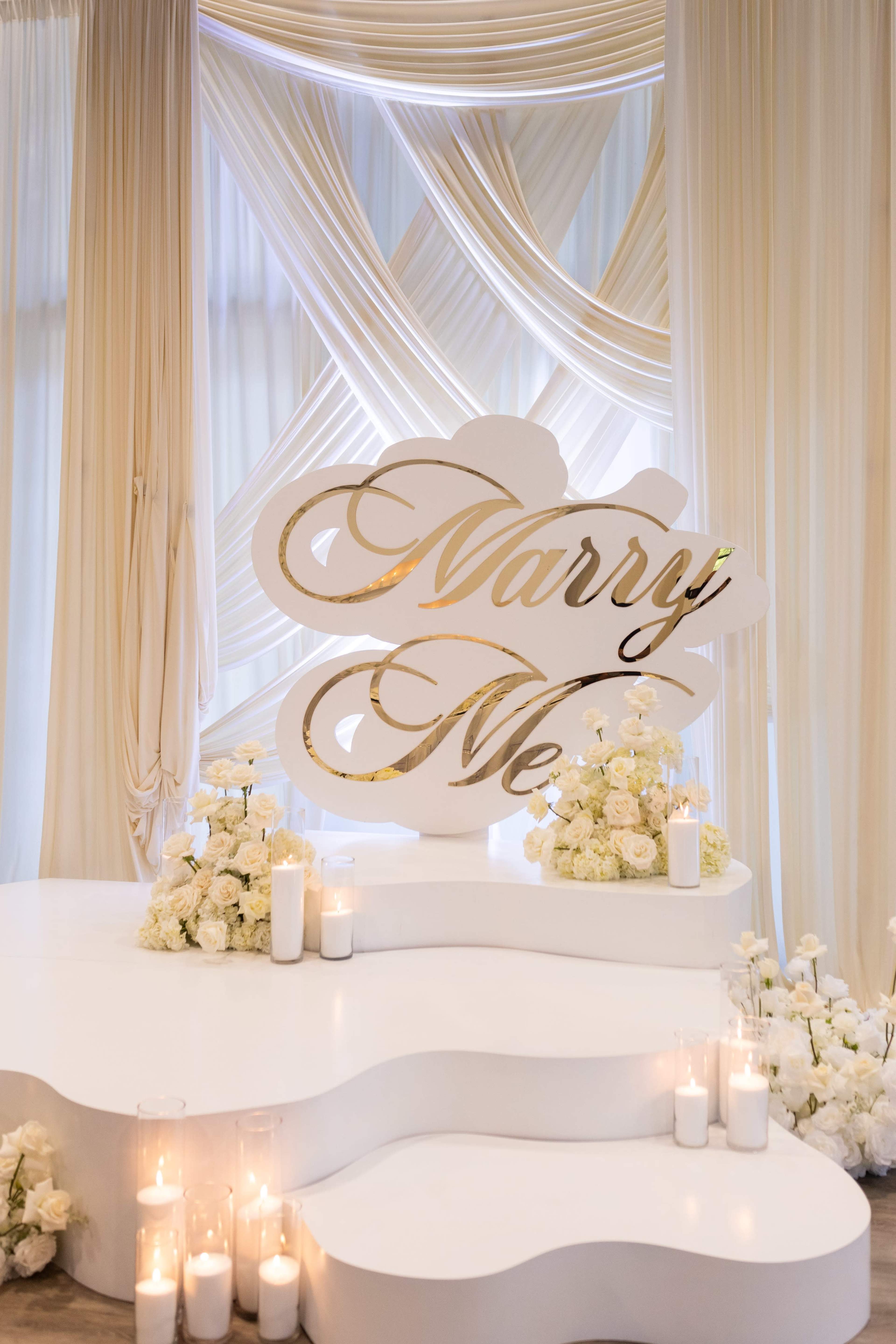 A large white sign with the words "Marry Me" is prominently displayed against a backdrop of draped fabric and floral arrangements, with candles arranged around it.