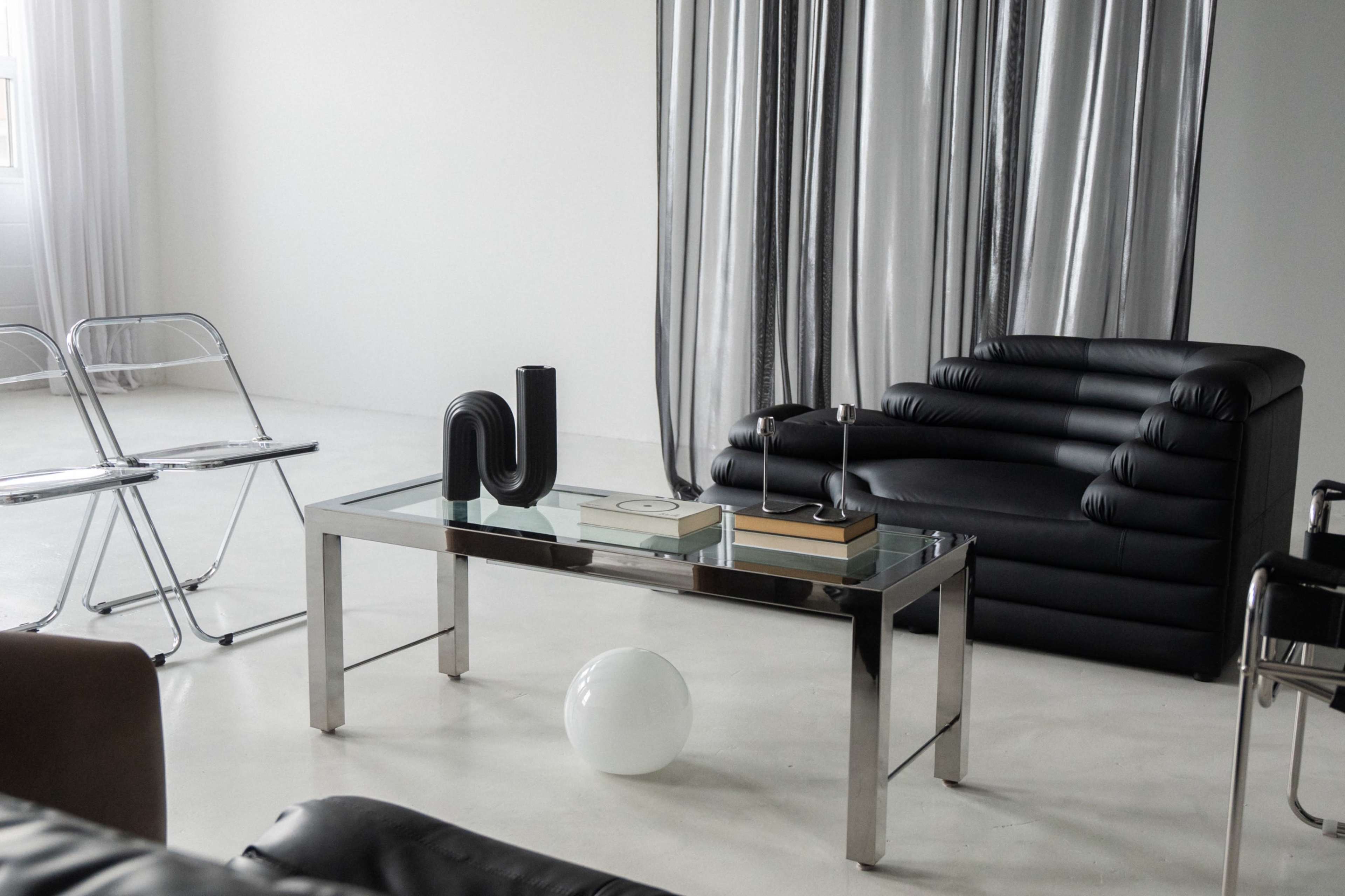 The image shows a modern living room with a black leather sofa, a glass coffee table featuring books and decorative items, chrome chairs, and silver drapes.
